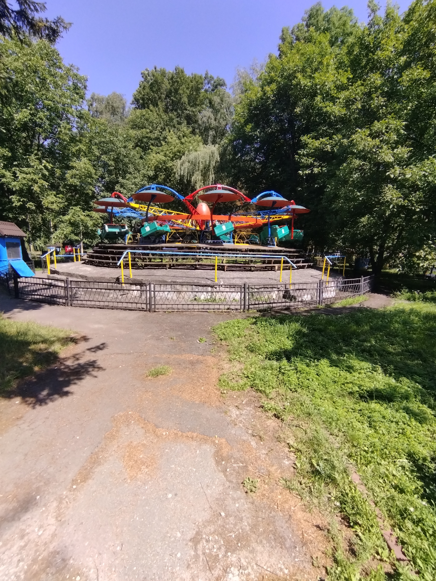 The image captures a colorful amusement park ride, possibly a carousel or a similar spinning attraction, situated outdoors in a park-like setting. The ride features several individual carriages, each adorned with a vibrant umbrella-like canopy in shades of red, orange, and blue. The carriages themselves are painted green and appear to be empty. The ride is surrounded by a dark-colored fence with a crisscross pattern, supported by yellow posts. Beyond the ride, a dense arrangement of lush green trees forms a backdrop, suggesting a natural or park environment. A small blue structure with a blue slide is visible to the left of the ride. The ground in the foreground is a mix of dirt and sparse patches of grass. The sky above is clear and blue. The overall impression is that of a sunny day in a recreational area. This scene is consistent with an outdoor public space in Khmelnytskyi, Ukraine, given the characteristic green spaces and amusement attractions often found in such locations.