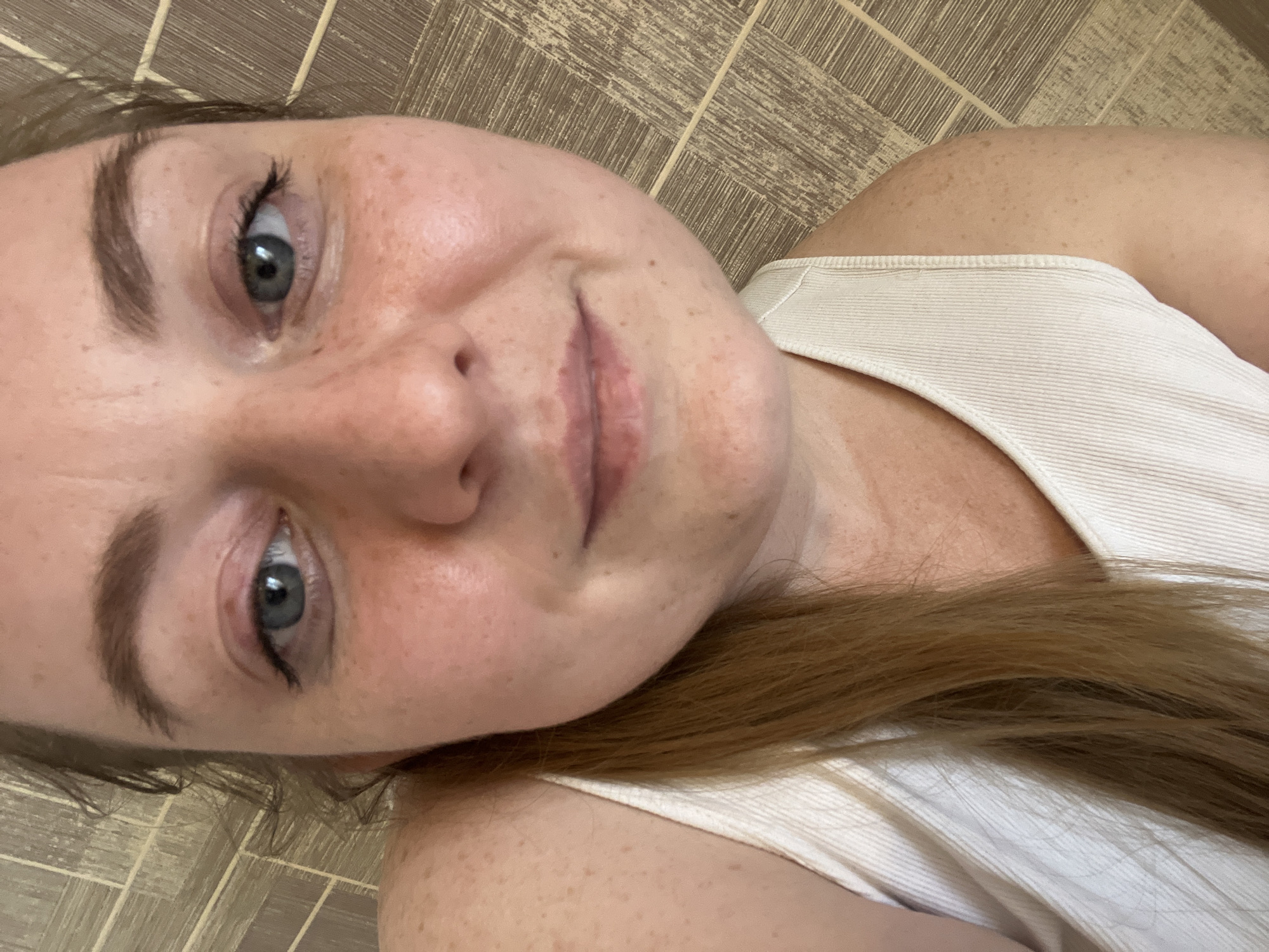 A close-up, rotated image depicts a fair-skinned individual with freckles, blue eyes, and light brown hair. The person is wearing a white ribbed tank top. The background consists of a brown tile floor. There is no indication of the location being Раздолье, Russia.