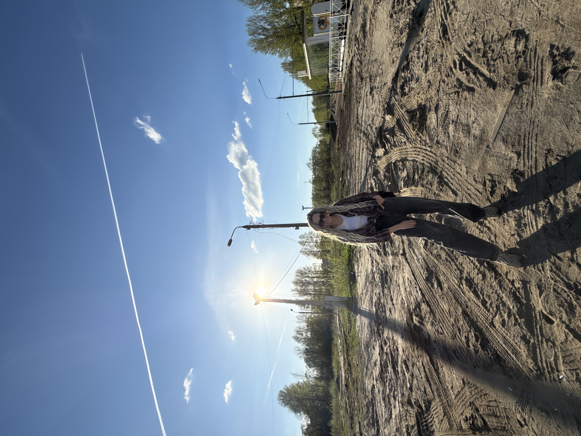 The image depicts an outdoor scene in what appears to be a disturbed or undeveloped area in or near Rusanovo, Russia. The setting is a sandy or dirt-covered ground showing tracks of tires and footprints. A young adult female stands on this ground wearing sunglasses, a white top, a plaid jacket, and dark pants. Behind the female, there are utility poles, trees, a bus stop, and possible train tracks. The sky is blue with a contrail and some clouds. The sun is visible and casts a strong glare into the camera.