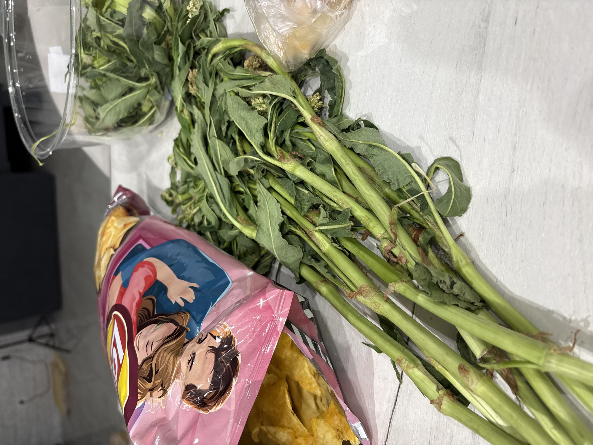 The image shows a still life composition on a white surface with wood grain texture. A partially open bag of Lays potato chips, featuring an image of two embracing people on the packaging, is positioned on the left side of the frame. To the right of the chip bag, there is a bunch of green leafy vegetables with thick green stalks. The vegetables appear to be mature, with some flowering buds visible. In the upper left corner, there is a clear plastic container filled with similar green leafy vegetables. A clear plastic bag sits above the bunch of leafy vegetables. The setting and location of the scene are unspecified.