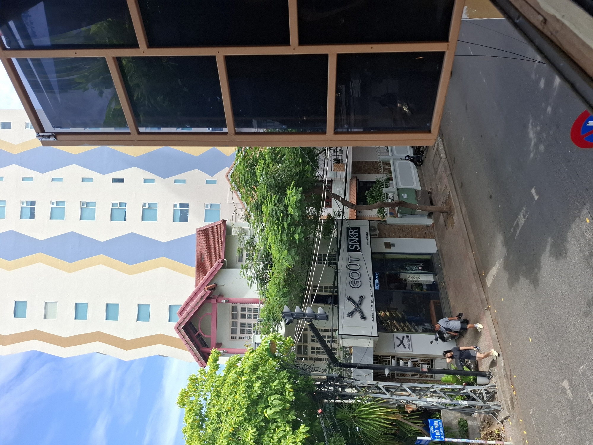 In the city of Nha Trang, Vietnam, this image captures a street scene from an elevated perspective. A tall, modern building with a distinctive zigzag pattern of beige, white, and blue stands prominently in the background. In the middle ground, a smaller, pink-roofed house with white walls and balconies is nestled amongst lush green trees. A storefront with the name "GOUT SNKR" is visible below the house, featuring a large window displaying shoes.
In the foreground, a paved street runs along the sidewalk where two people are walking. A woman in a black t-shirt and shorts walks ahead of a man in a grey t-shirt and shorts, who is carrying a pair of shoes. To the left, a traffic light and a street sign are mounted on a utility pole. The street is marked with white lines, and a blue and red circular traffic sign is visible near the bottom right of the frame. The overall scene depicts a bright, sunny day in an urban environment.