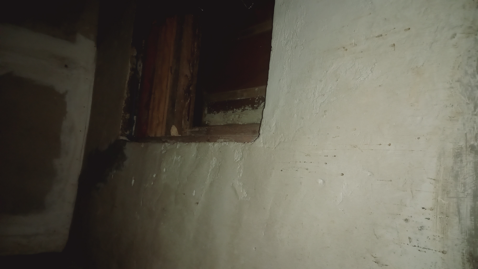 This image depicts a close-up view of a rough, weathered wall with an opening that reveals wooden structural elements within. The wall appears to be made of concrete or stucco, exhibiting signs of age and damage, with patches of peeling paint or plaster. The opening, which is dark and indistinct, suggests a structural cavity or perhaps an unfinished window frame. The overall texture of the wall is uneven, with visible marks, stains, and small imperfections that contribute to a sense of neglect or disuse. The lighting is dim, casting shadows and highlighting the textured surface of the wall. The scene is static, with no discernible activity taking place. The context of Leuwisadeng, Indonesia, suggests this might be an exterior or interior wall of a building in that region. No people, animals, or text are visible in the image.