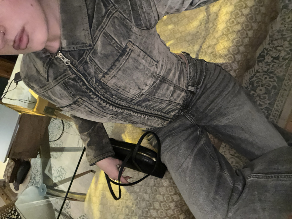 The image captures an individual in an indoor setting, observed from an elevated, slightly oblique perspective. The subject is seated or reclining on a light brown patterned upholstered surface. They wear a fully zipped, distressed grey denim jacket with a collar and chest pockets, paired with grey denim jeans. The subject's left hand, adorned with a gold-colored ring on the ring finger, holds a black shoulder bag with a gold zipper. A portion of the subject's face is visible at the upper right, showing the mouth with dark lip color and a nose ring in the right nostril. In the background to the left, a glass-top table with a silver base is visible, upon which an open silver-colored laptop, a wooden box, and a dark rectangular object are placed. Electrical wires are present behind the table. A darker patterned fabric is visible beneath the subject's right leg and extends to the lower left of the frame. The lighting within the scene appears warm. The specific location within North Hertfordshire, United Kingdom, cannot be determined from the visual information provided.