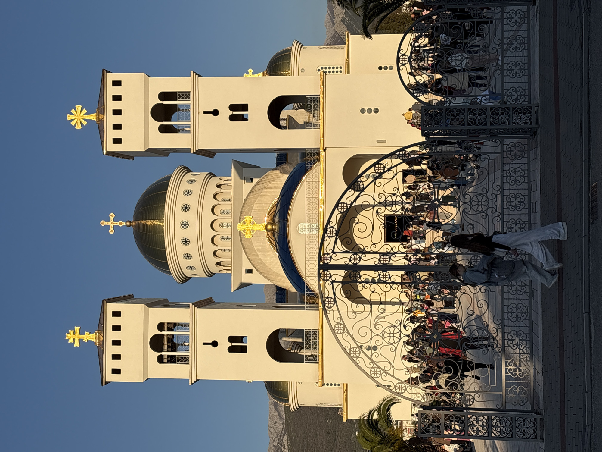 The image depicts a modern-style Orthodox church in the city of Bar, Montenegro. The architecture is characterized by two bell towers flanking a central dome, all painted in a light beige color with golden accents. Each tower is topped with a golden cross.
A wrought-iron gate separates the church from the public area. A crowd of people is seen, gathered on the other side of the gate, suggesting a gathering or event. Two people are visible in the foreground, walking toward the church entrance.
The background reveals a clear blue sky and distant mountains. A palm tree is partially visible at the bottom left corner of the image. The photograph appears to be taken during daylight hours, with sunlight casting shadows on the building.