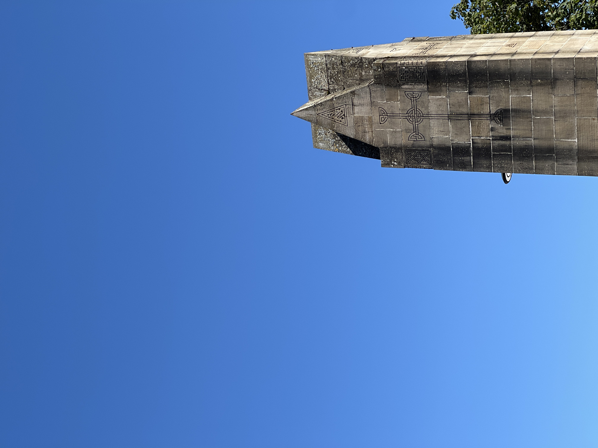 The image captures a close-up, upward view of a stone tower against a clear blue sky. The tower, made of large, roughly hewn stone blocks, is topped with a pointed, triangular section. A Celtic cross motif is carved into the stone on one face of the tower, along with geometric patterns on either side. The stone appears weathered and somewhat mossy in places. To the right of the tower, the edge of a green tree canopy is visible. A small, dark object, possibly a light fixture, is attached to the side of the tower in the lower left portion of the frame. The scene is devoid of people. The tower's design suggests it may be a monument or part of a historical structure. Without further information, its specific location within Donegal, Ireland, cannot be determined from the image alone.