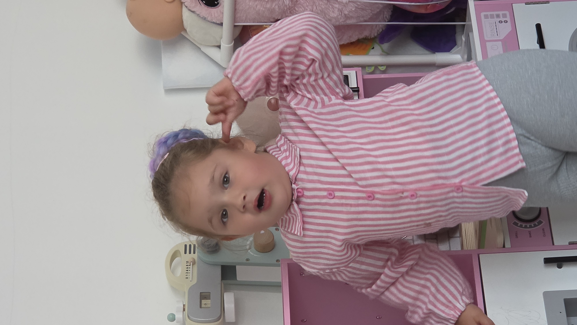 A young girl with light skin and blue eyes, wearing a pink and white striped pajama top and gray leggings, points to her left ear with her right index finger. She has light brown hair with purple and blue highlights gathered in a headband. The girl is positioned in front of a pink and white toy kitchen, which includes a play oven, a sink, and various accessories. Soft toys are visible in the background. The location is an indoor domestic setting, but there is no indication of it being in London.