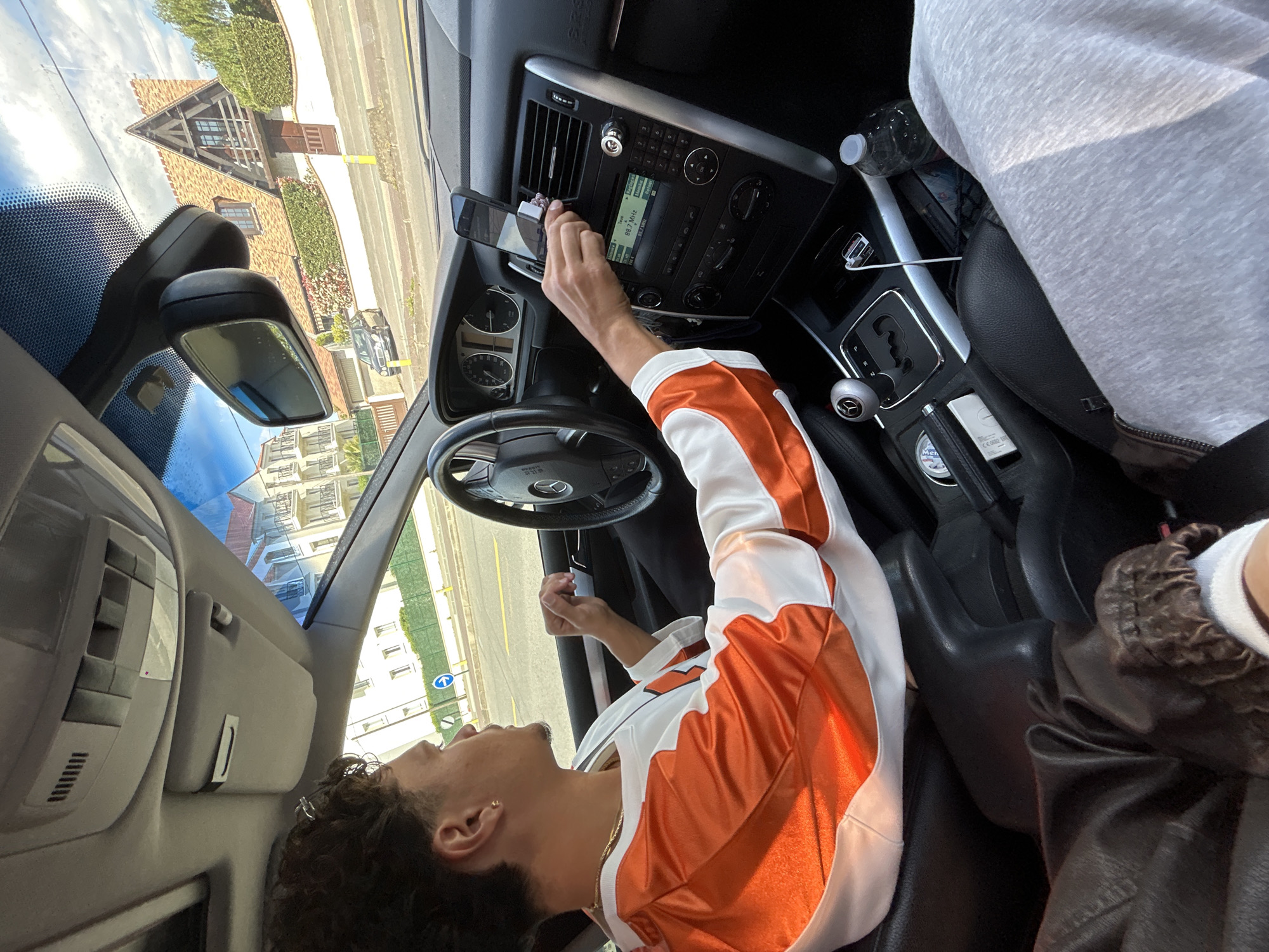 The interior of a vehicle is depicted in the image. A young male with dark curly hair, wearing a white and orange jacket, is in the driver's seat, his hand reaching towards the dashboard. A mobile phone is attached to the air vent. The car's dashboard features a stereo system. A second person, whose body is partially visible, is seated in the passenger seat wearing a grey sweatshirt. A water bottle is placed to the side of the gear stick. Outside the vehicle, buildings and a street are visible. The architecture and street signage suggest the location is Noisy-le-Grand, France.