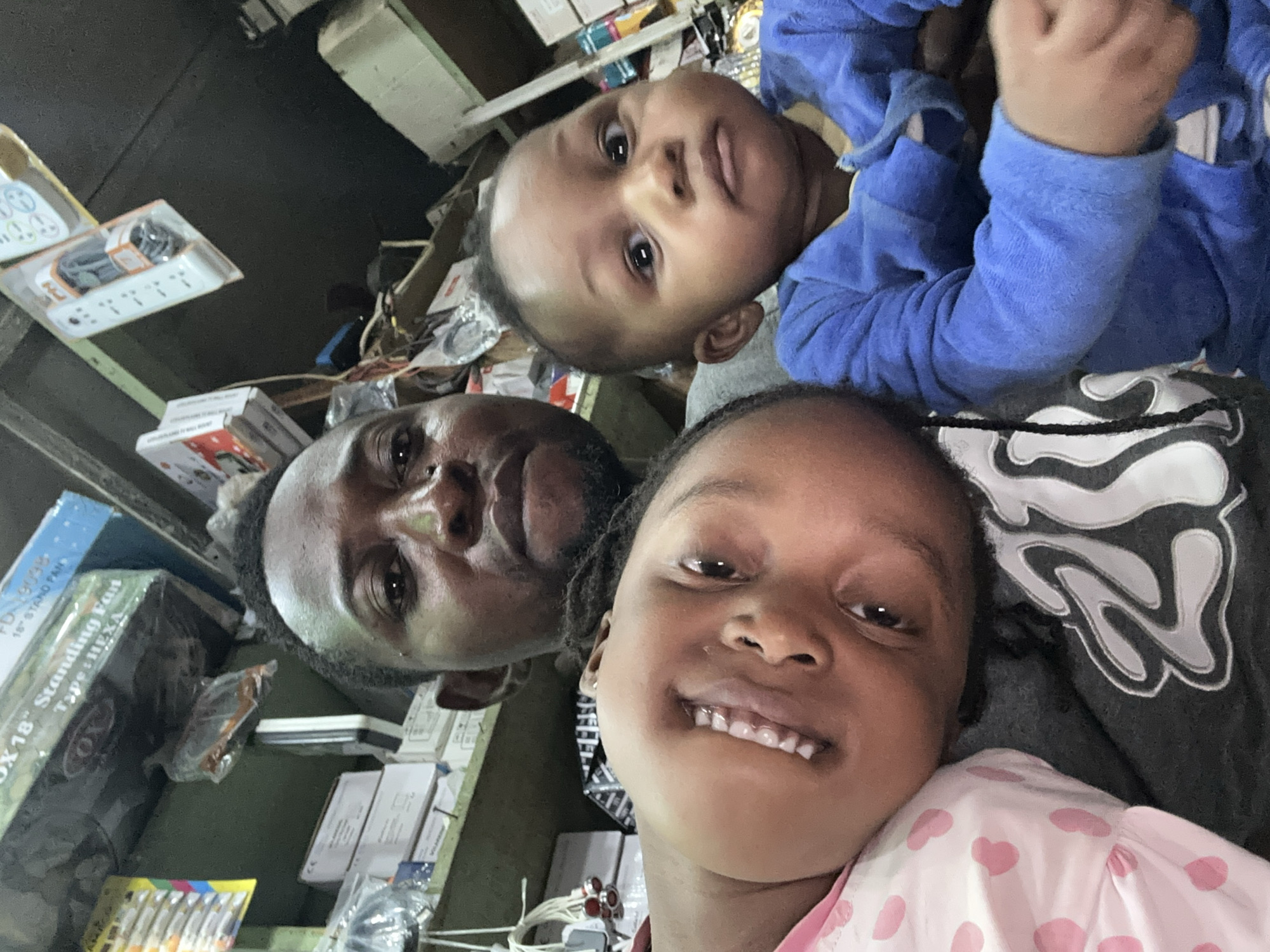 A selfie captures an adult male with two children in what appears to be a small retail shop in Narayi, Nigeria. The adult male, positioned centrally at the top, has dark skin, short dark hair, a slight beard, and a neutral expression as he looks directly into the camera. A young girl with dark skin and dark hair is on the bottom-left, resting her head against the man's shoulder/chest, smiling broadly with her upper and lower front teeth visible. She wears a pink and white polka-dot top. An infant with dark skin, wide eyes, and a neutral expression is held by the man on the bottom-right, wearing a blue hooded fuzzy jumpsuit.
The background environment consists of cluttered shelves stocked with various items. On the top left shelf, a cardboard box is visible with text "OX 18" Standing Fan Type: HEXA" and another indicating "FD-1909B 16" STAND FAN". Below this, more white boxes, a clear plastic bag with orange contents, and a white power strip are visible. On the right side, another white multi-outlet power strip hangs from a shelf, with additional boxes, wires, and general merchandise filling the space, characteristic of a small electronics or general goods store.