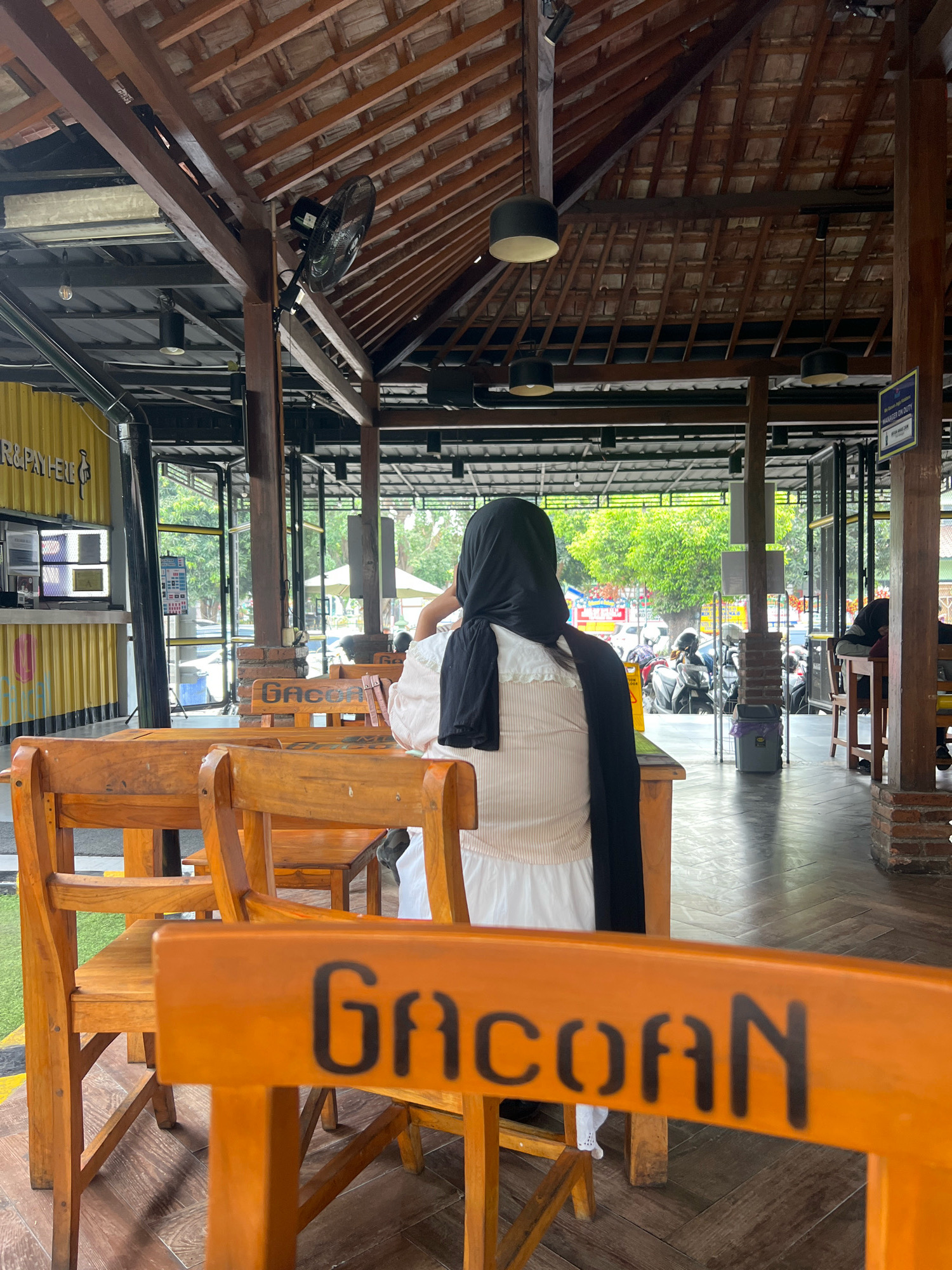 A woman wearing a black hijab and a beige and white outfit sits facing away from the camera in an outdoor covered dining area. She is seated at a wooden table, with her right hand touching her face. In the foreground, a wooden chair with the word "GACOAN" stenciled on it in black is visible. The setting appears to be a restaurant or cafe with a wooden roof structure, exposed beams, and tiled roofing. In the background, several motorcycles are parked, and trees and a yellow building are visible through open-air entrances. The flooring is made of wood planks. The scene is well-lit, suggesting it is daytime. The establishment is likely located in Yogyakarta, Indonesia, given the presence of the word "GACOAN", which is a popular food chain in that region.