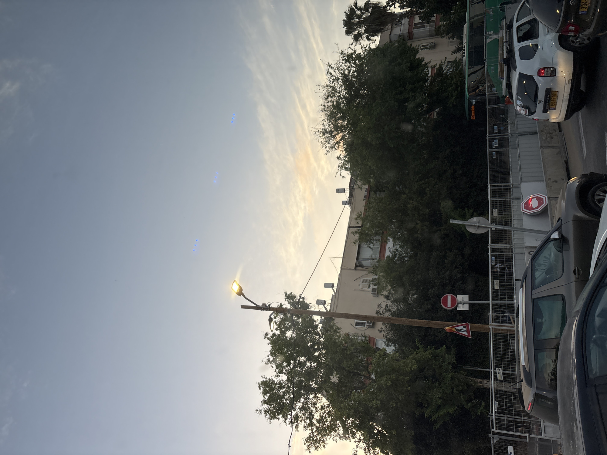 The image depicts a street scene in Tel Aviv, Israel, at dusk. The sky is a pale blue with streaks of orange and pink clouds, indicating the setting sun. A street lamp is illuminated, casting a warm glow. Several trees, some with dense green foliage and one with palm fronds, are visible. In the background, a multi-story building with balconies and air conditioning units is partially obscured by the trees.
The street level shows a traffic jam. Several cars are present, including a grey SUV, a white Dacia Duster with a license plate reading "185-55-10", and the rear of a dark-colored car. A green bus with "135 Central Station" printed on its side is also in the traffic. Construction barriers with metal fencing are set up along the side of the road.
Several traffic signs are visible: a triangular sign with a pedestrian symbol and a fallen figure, a circular red and white "no entry" sign, and a circular red sign with a white horizontal bar. In the sky, three sets of faint blue lights are visible, arranged in triangular patterns. It is unclear what these lights represent. The overall atmosphere is that of a typical urban evening commute.