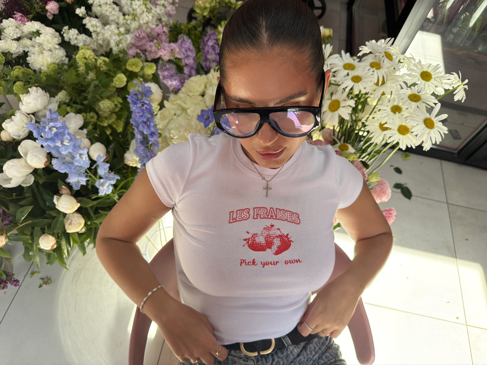 A woman with her hair pulled back stands or sits among a large bouquet of flowers, including daisies, peonies, and delphiniums. She wears oversized black-rimmed glasses, a white T-shirt with the words "LES FRAISES" and "Pick your own" printed on it, a thin gold necklace with a cross pendant, a thin pearl bracelet on her left wrist, and a black belt. She has rings on the fingers of both hands and is adjusting her belt. The location is possibly Krasnoyarsk, Russia based on metadata.