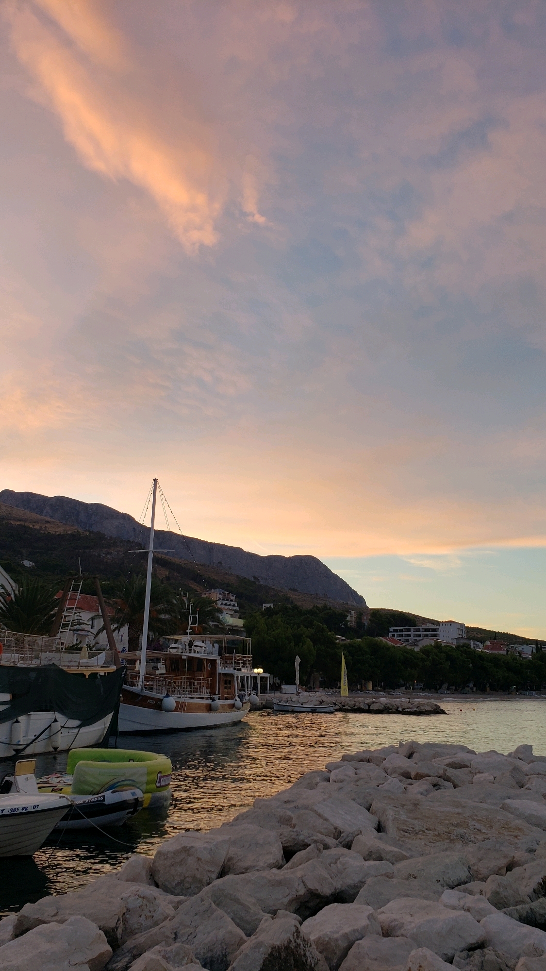 This image depicts a tranquil waterfront scene in Općina Tučepi, Croatia, during a sunset. The sky is a soft gradient of pink, orange, and pale blue, with scattered clouds. In the foreground, a rocky breakwater lines the edge of the water, with several boats moored nearby. One boat, a larger wooden vessel with white railings and a mast, is prominently featured. Smaller inflatable boats and a dinghy are also visible. The water reflects the warm hues of the sky. In the background, a mountainous landscape with lush green trees and buildings is illuminated by the fading sunlight. A few palm trees are also present along the coastline. The overall atmosphere is serene and picturesque. There are no people visible in the image.