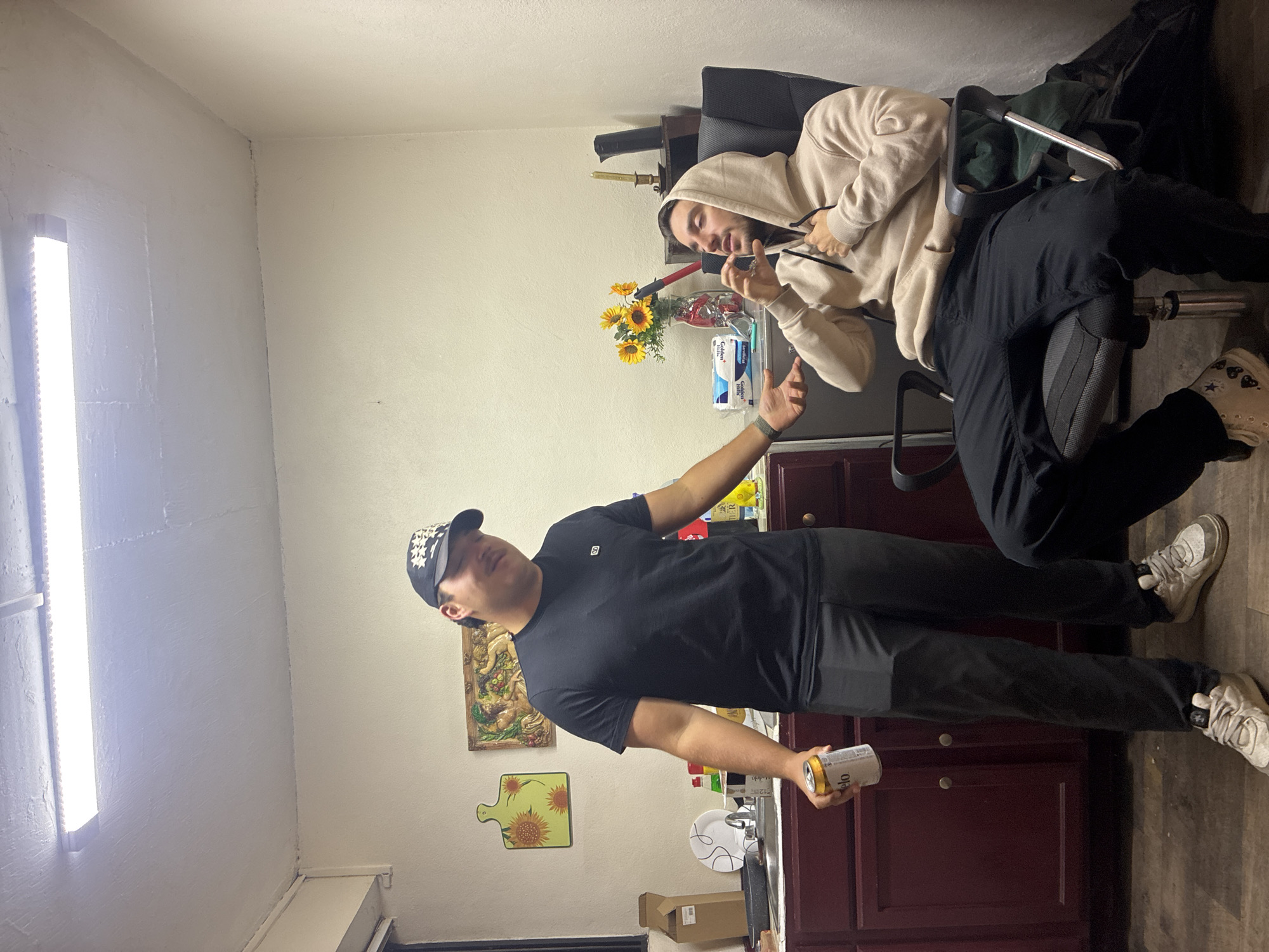 A fluorescent light fixture illuminates a narrow room in Huixquilucan de Degollado, Mexico. The walls are painted white. A man wearing a black t-shirt, dark gray pants, a black baseball cap with white floral designs, and white sneakers stands in the center of the room, holding a can of Modelo beer in his right hand. A second man sits on an office chair behind the first, wearing a tan hooded sweatshirt, black pants, and white sneakers. The second man is holding a red-handled object. The room features dark red cabinets, a sink, and a counter. On the counter are a vase of sunflowers, a candle, a box of tissues, and other miscellaneous items. On the wall above the counter hang a framed painting of fruit and a cutting board shaped like a sunflower. A black office chair is also visible.