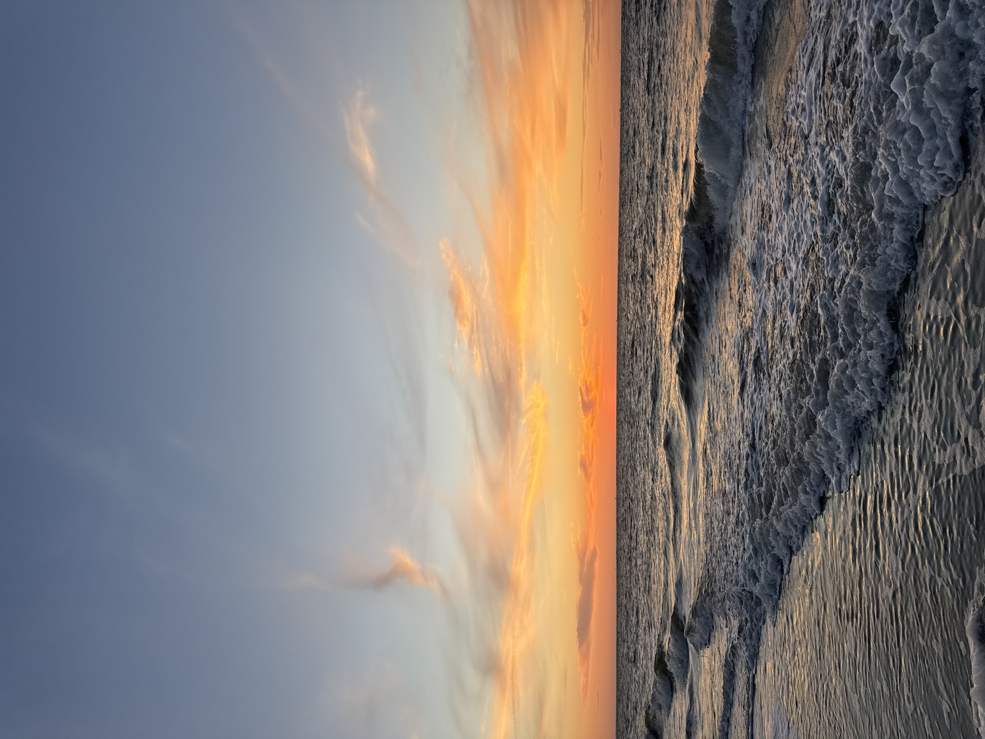A wide-angle photograph captures the tranquil scene of the North Sea at sunset on the island of Langeoog, Germany. The horizon is marked by a gradient of warm colors, transitioning from vibrant orange near the water to softer pinks and yellows in the sky, with streaks of pale blue and wispy clouds above. Gentle waves roll towards the shore, their white foam creating delicate patterns on the dark water. The water itself reflects the warm hues of the sunset, creating a shimmering effect. The foreground is the sandy beach, wet from the receding tide and reflecting the sky's colors. No people or man-made objects are visible, emphasizing the natural beauty of the coastal landscape.