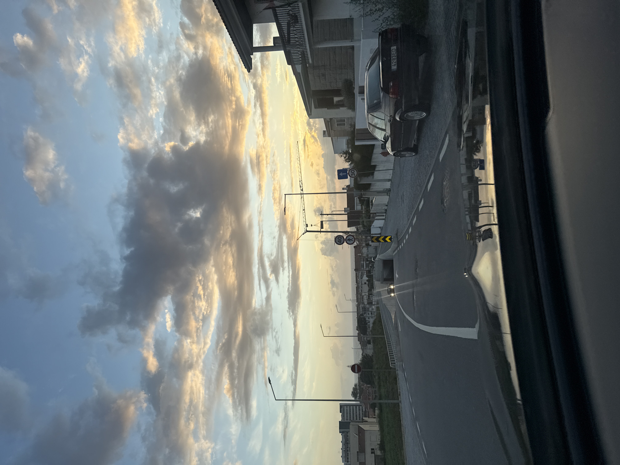 Captured from within a vehicle, the image depicts a streetscape in Vila Nova de Gaia, Portugal, under a dramatic sky of layered clouds illuminated by a setting sun. The view is oriented along a road, with adjacent buildings and parked cars lining the street. Road signs are visible. A streetlamp casts a warm glow, suggesting dusk or early evening. The image suggests a moment of urban life captured from a moving or stationary vehicle.