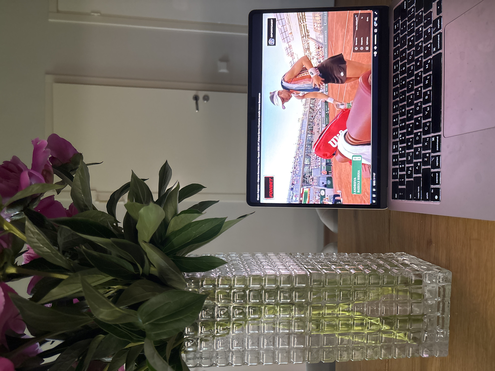 Here is a matter-of-fact description of the scene:
A laptop shows a tennis match on its screen, specifically a match between Mirra Andreeva and Marina Bassols Ribera at Roland Garros 2026. The screen also displays the Sportcast logo and the Fonbet logo. The laptop keyboard is visible and is in a Cyrillic layout. To the left of the laptop is a rectangular glass vase filled with water and a large bouquet of pink flowers and green leaves. The vase and laptop sit on a wooden surface. Behind them is a white wall with a door and a door handle.