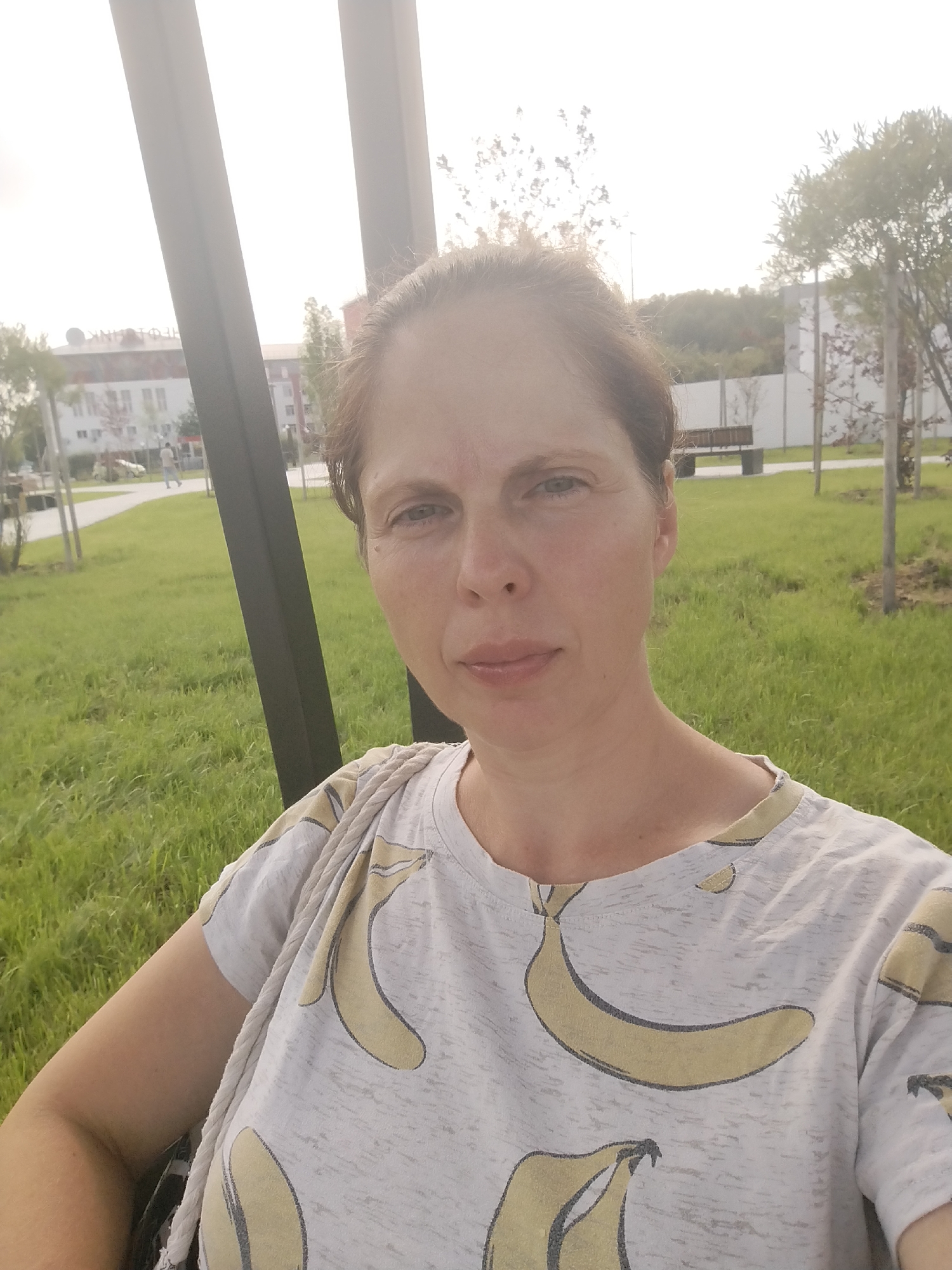 The image is a medium-shot selfie of a woman with brown hair and fair skin. She is wearing a light-colored t-shirt with a banana pattern. Her expression is neutral. The background shows a park with green grass, trees, and a building in the distance. There are also some dark vertical structures in the foreground, possibly part of a swing set or park bench. The sky is bright and appears overcast or hazy. The location is identified as a park in the city of Tyumen, Russia.