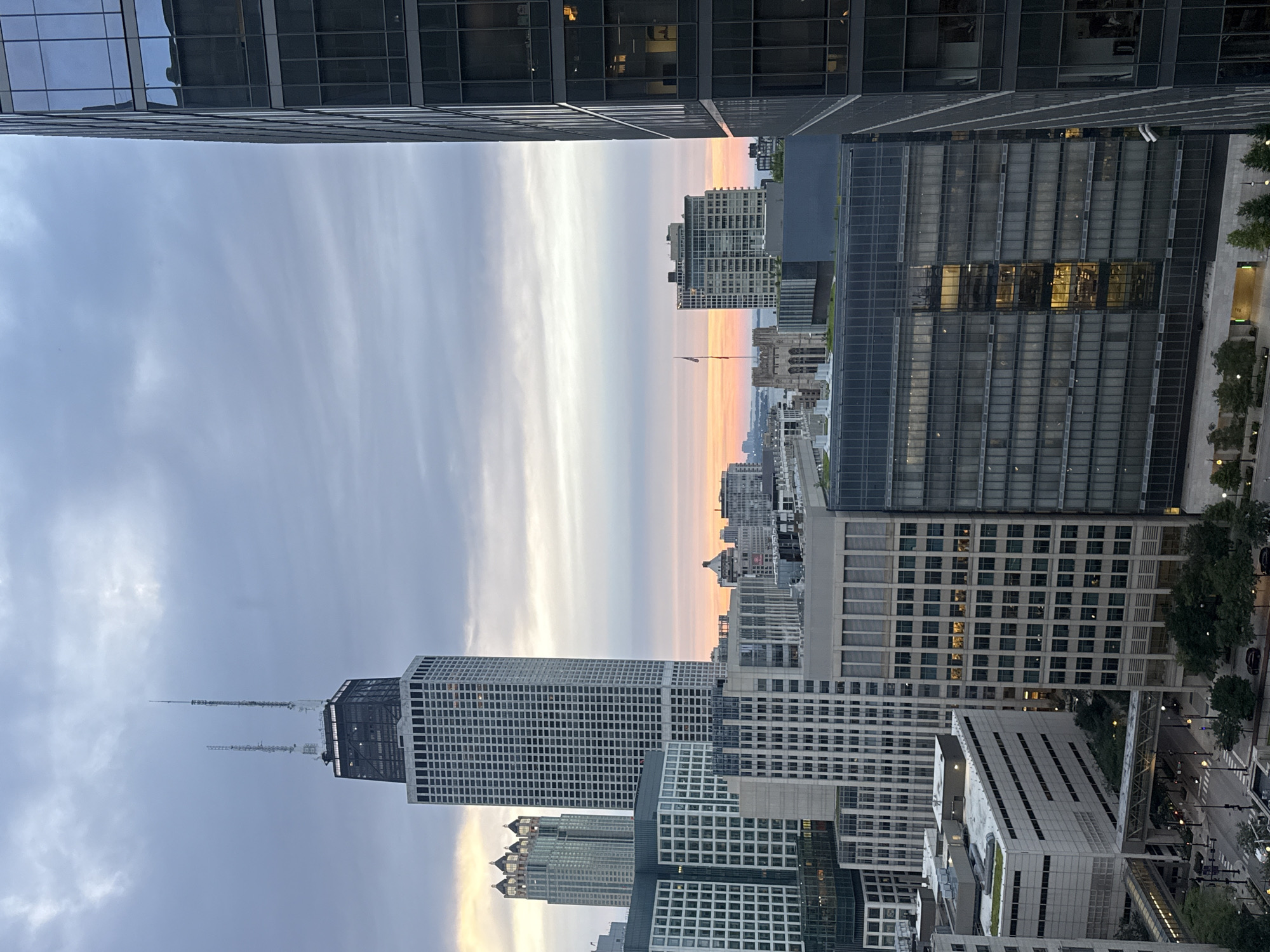 This is a photograph of the Chicago skyline taken at dusk, with a view towards the east over Lake Michigan. The iconic John Hancock Center is prominently featured in the middle ground, its distinctive structure silhouetted against the soft hues of the setting sun. The sky is a canvas of grays and pinks, with wispy clouds suggesting a transition from day to night.
Surrounding the Hancock Center are numerous other skyscrapers, their glass facades reflecting the fading light. To the right, a modern, dark glass skyscraper dominates the foreground, its sheer verticality contrasting with the more complex architecture of the older buildings. The buildings are densely packed, indicative of a major metropolitan area.
Below the skyscrapers, the street level is visible, with streetlights beginning to illuminate and a few cars moving along the road. Trees line some of the sidewalks, adding a touch of greenery to the urban landscape. Skybridges connect some of the buildings, further emphasizing the interconnectedness of the city.
In the distance, across the water, the lights of the far shore are just beginning to twinkle, hinting at the vastness of Lake Michigan and the spread of the urban area. The overall impression is one of a bustling metropolis at the close of a day, with the architecture of Chicago standing as a testament to its economic and cultural significance.