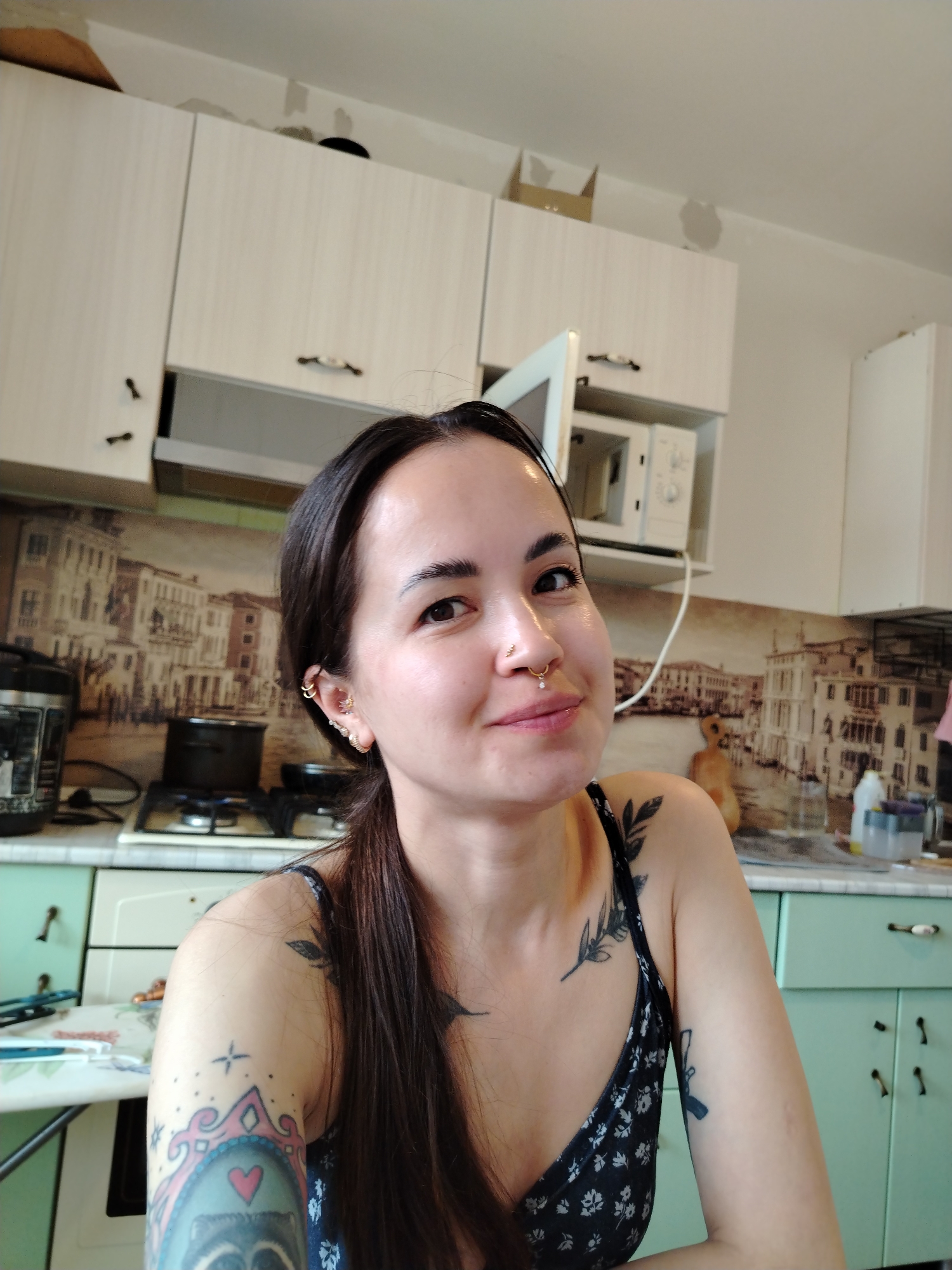 A woman is pictured indoors, presumably in a kitchen. She has dark brown hair pulled back into a ponytail, fair skin, and multiple piercings in her left ear and nose. She is wearing a dark-colored tank top with a floral pattern and has several tattoos on her arms and shoulders.
The background reveals a kitchen setting. The kitchen cabinets are light green and white with black handles. A microwave is mounted under one of the cabinets. The backsplash features a photographic image of a European city landscape, which is possibly Venice. A stove is visible with a black pot on it. The overall environment appears domestic and lived-in.
The image does not contain information pertaining to the city of Noviy, Russia.