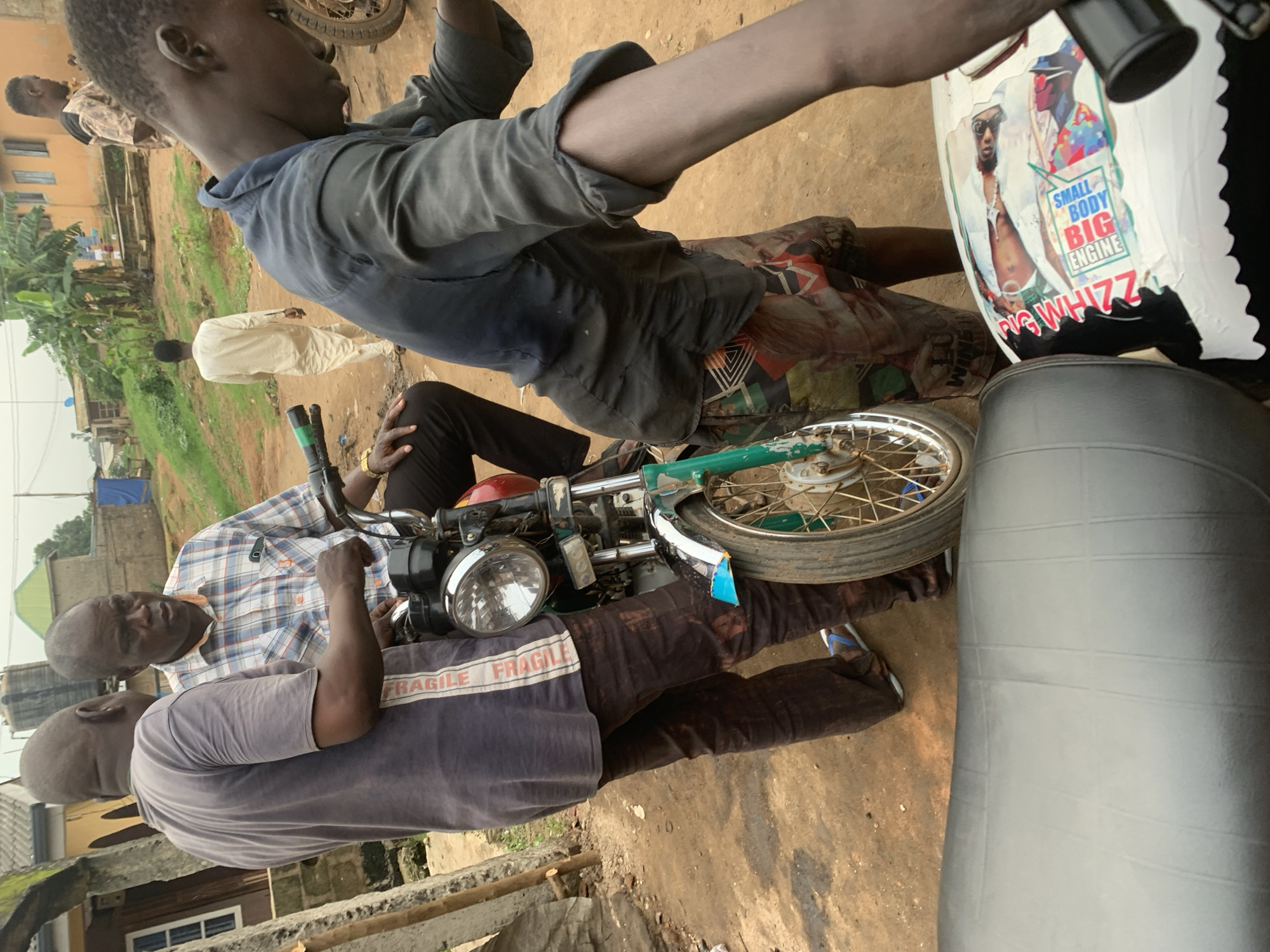 In Shagamu, Nigeria, three men are gathered around a motorcycle. One man, wearing a gray t-shirt with "FRAGILE" printed vertically on the side and dark brown pants, stands on the left, facing away from the camera and towards the other two men. Another man, dressed in a plaid shirt and dark pants, sits on the motorcycle, looking towards the man in the gray t-shirt. A third man, wearing a dark blue, soiled shirt and patterned shorts, is on the right, leaning over the motorcycle. In the background, a fourth man in a light-colored outfit walks away from the camera, and buildings, vegetation, and a water tank are visible. The ground is dirt. The motorcycle has a green fender and a white fuel tank adorned with stickers, including one with the text "SMALL BODY BIG ENGINE." The scene appears to be in an outdoor, possibly semi-urban or rural, area.