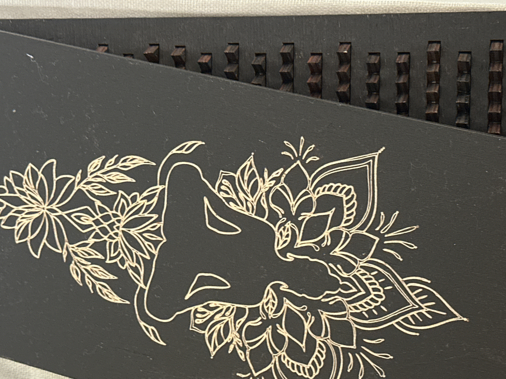 The image shows a close-up of a black surface with a gold line drawing of a floral design and a silhouette of an elephant's head. To the right of the black surface is a thin, dark-colored object with a series of small, dark wooden protrusions arranged in rows. The background is a textured off-white material. There are no people or any indication of a specific location in Nevinnomyssk, Russia.
