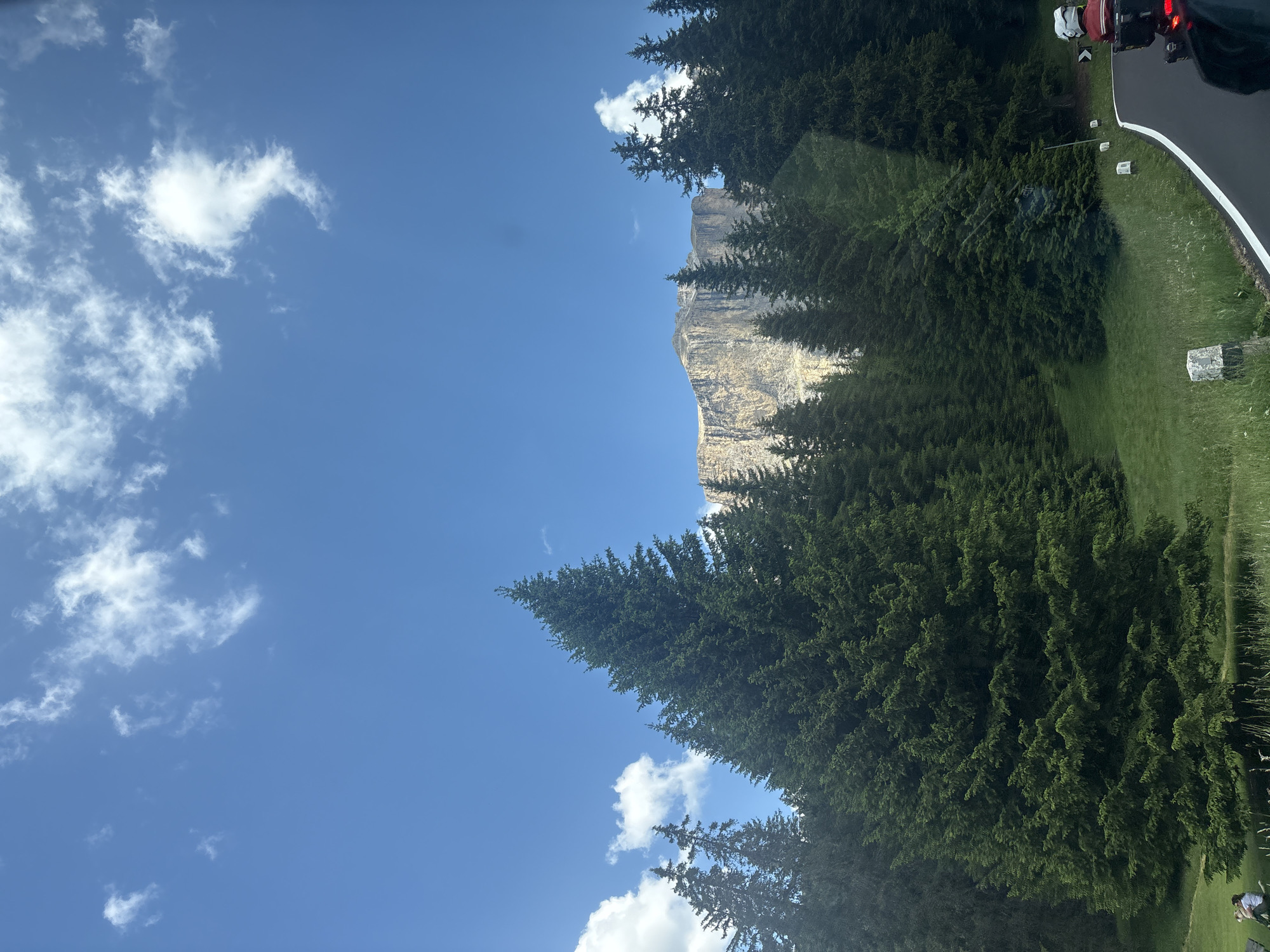 The image depicts a serene mountain landscape under a bright blue sky dotted with white clouds. In the foreground, a winding asphalt road curves to the right, bordered by a grassy verge and small, white-topped marker posts. To the left of the road, a dense forest of dark green evergreen trees fills much of the frame, with a large, prominent fir tree in the middle ground. Behind the trees, a towering, light-colored mountain range with sheer cliffs dominates the background. A small section of the mountain appears to be illuminated by sunlight, highlighting its rugged texture. In the far lower-left corner, two people are seated on a bench amidst the grass, facing away from the camera. On the right side of the road, a motorcycle with a rider wearing a white helmet and red jacket is visible, with its taillight illuminated. The overall scene conveys a sense of natural beauty and a peaceful, outdoor setting.
