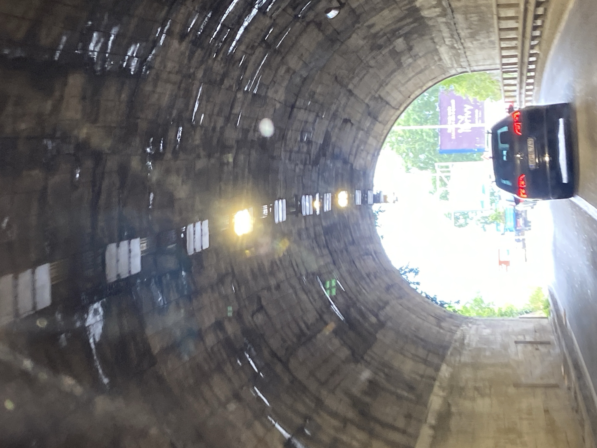 The image presents a view from inside a dark, concrete road tunnel, looking towards its bright exit. The tunnel walls are textured and appear damp or wet, with visible streaks and reflections from moisture. Rows of bright lights are installed along the upper sections of the tunnel, some of which are intensely glowing, creating lens flare.
Ahead, a black car, resembling an SUV or hatchback, is positioned near the tunnel's opening, driving forward. Its red taillights are illuminated. A partially visible license plate reads "BD BH 078", and a white, vertical marking or sticker is on its rear window.
Beyond the tunnel exit, bright daylight illuminates lush green foliage and what appears to be a large, purple billboard. The text on the billboard is not entirely legible due to distance and obstruction, but letters such as "A&L" or similar can be vaguely discerned in white. The scene depicts a daytime journey through a tunnel, suggesting an approach to an outdoor area. The overall perspective is from inside a vehicle, slightly tilted.