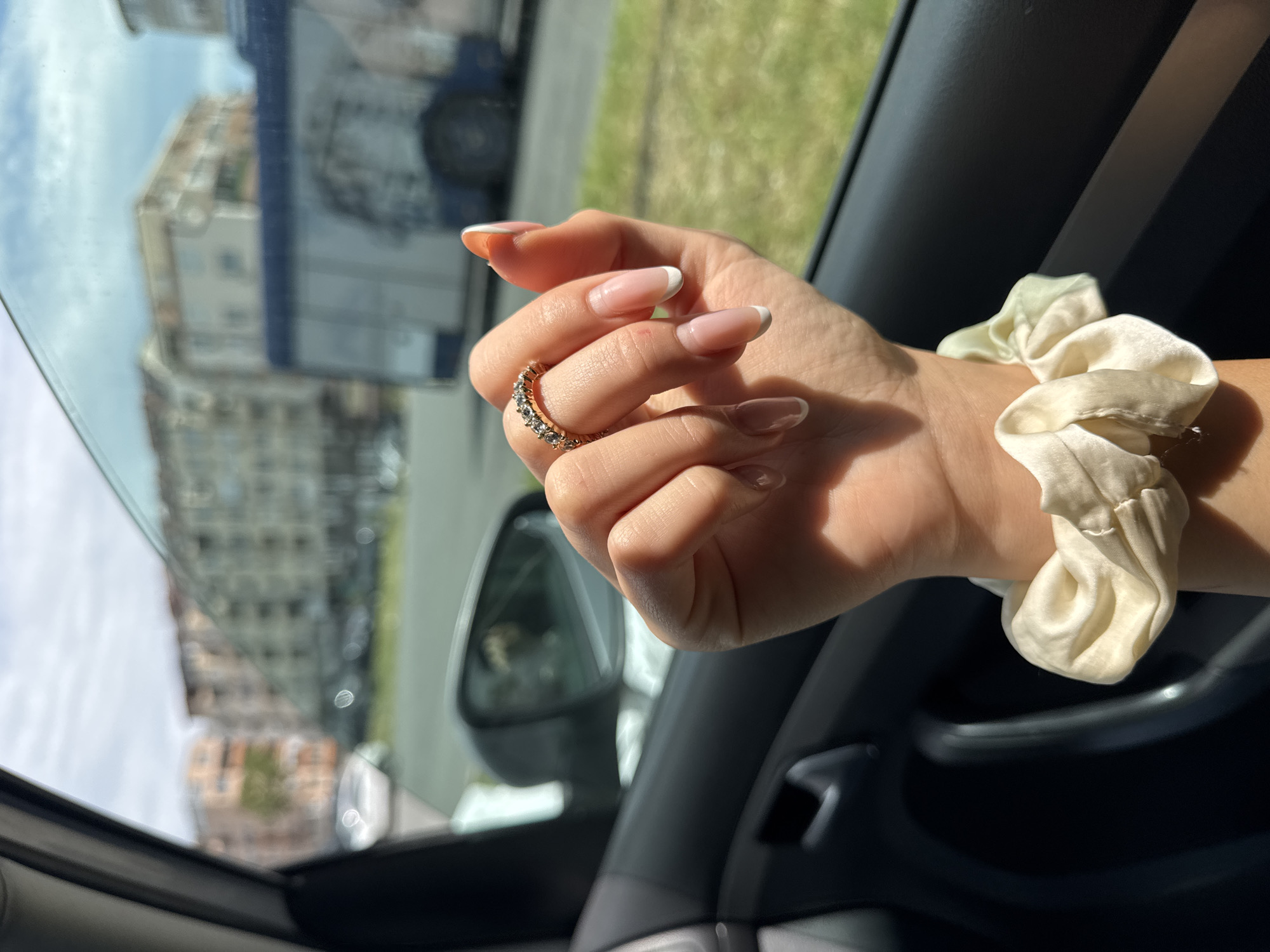 The image shows a person's hand adorned with a gold ring, resting on the arm of a car door. The hand has manicured nails with a white French tip design, and the wrist is decorated with a scrunchie. The background is blurred, but it is possible to discern a building and vegetation seen through the open car window. Based on the architectural style of the building and the presence of greenery, the location could be in Burgas, Bulgaria. The scene appears to be taken from inside a vehicle.