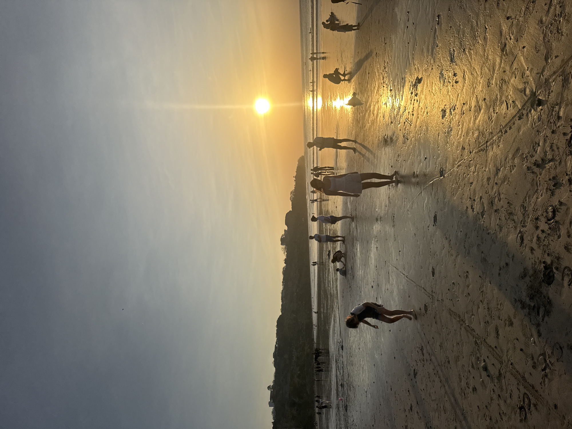 The image depicts a beach scene in Kuta Selatan, Indonesia, at sunset. The sun is setting over the ocean, casting a warm, golden glow across the sand and water. Several people are visible on the beach, some walking, others sitting or playing in the sand. The shoreline is lined with people and a few structures are visible on a hill in the background. The wet sand reflects the colors of the sunset, creating a shimmering effect.