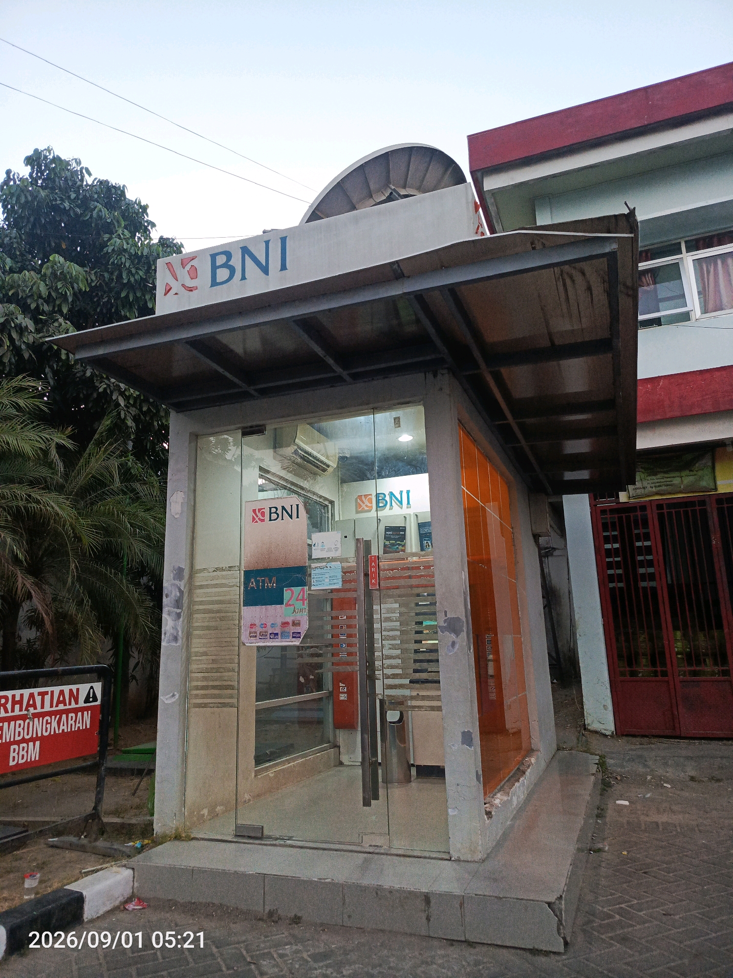 A glass-fronted ATM booth with a BNI logo is located in the city of Gladak Anyar, Indonesia. The booth is covered by a metal awning and is adjacent to a building with red and light blue siding. To the left of the ATM booth is a sign with the text "PERHATIAN PEMBONGKARAN BBM," indicating a warning about the demolition of BBM (likely referring to a fuel station or related facilities). Palm trees and other foliage are visible behind the booth. The ground is paved with bricks. The timestamp "2026/09/01 05:21" is superimposed on the lower left corner of the image.