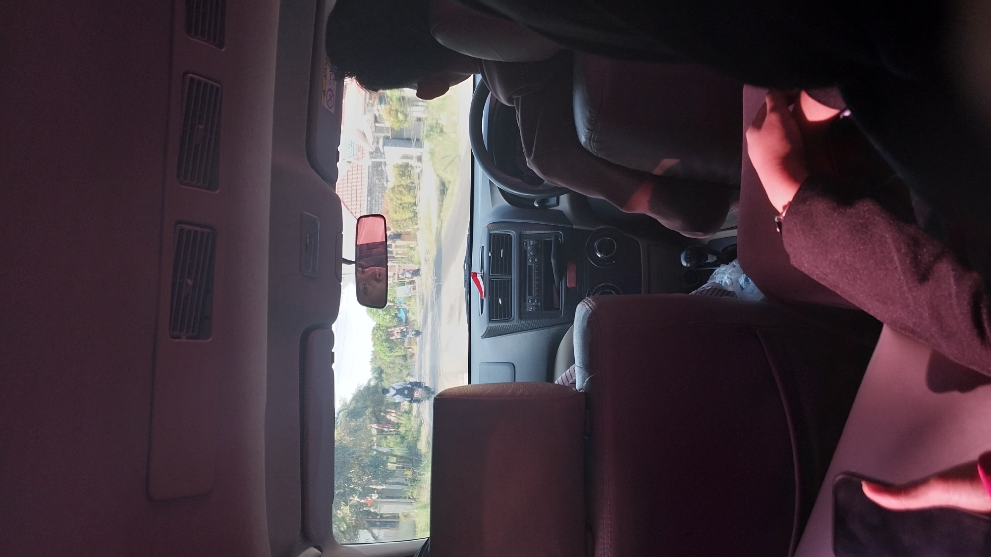 This is a medium shot from inside a vehicle, looking forward through the windshield. The perspective is from a passenger seat, with the back of the driver's head and shoulder visible in the right portion of the frame. The driver is wearing a brown hooded sweatshirt. In the foreground, the back of a passenger's head is visible on the left, with a portion of their arm and hand resting on their lap. The hand is holding a smartphone.
The vehicle's dashboard is clearly visible, featuring an audio system, air vents, and a gear shift. A small red and white flag is attached to the dashboard. Sunlight streams into the vehicle, casting shadows.
Outside the vehicle, the road stretches ahead, lined with trees and houses. Several motorcycles are visible on the road, some with riders. The scene appears to be a rural or suburban area, possibly in Southeast Asia, given the style of houses and motorcycles. The sky is bright, indicating daytime.