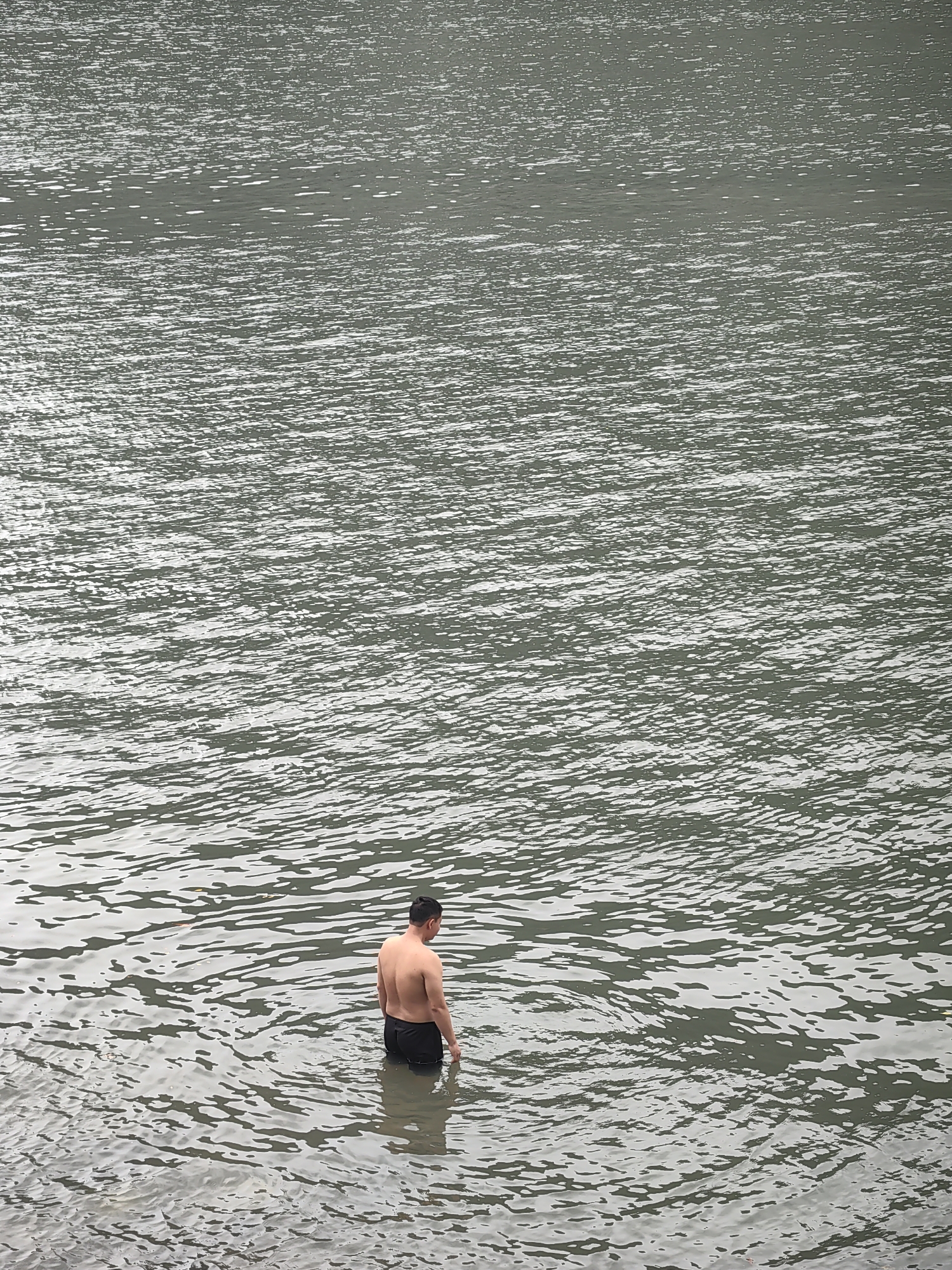A man stands in water in Hong Gai Ward, Vietnam. He is facing away from the viewer and the water is up to his waist. He is bare-chested and wearing dark shorts or swimwear. The water is gray and rippled. The background is filled with the same gray water. The lighting suggests it is an overcast day.