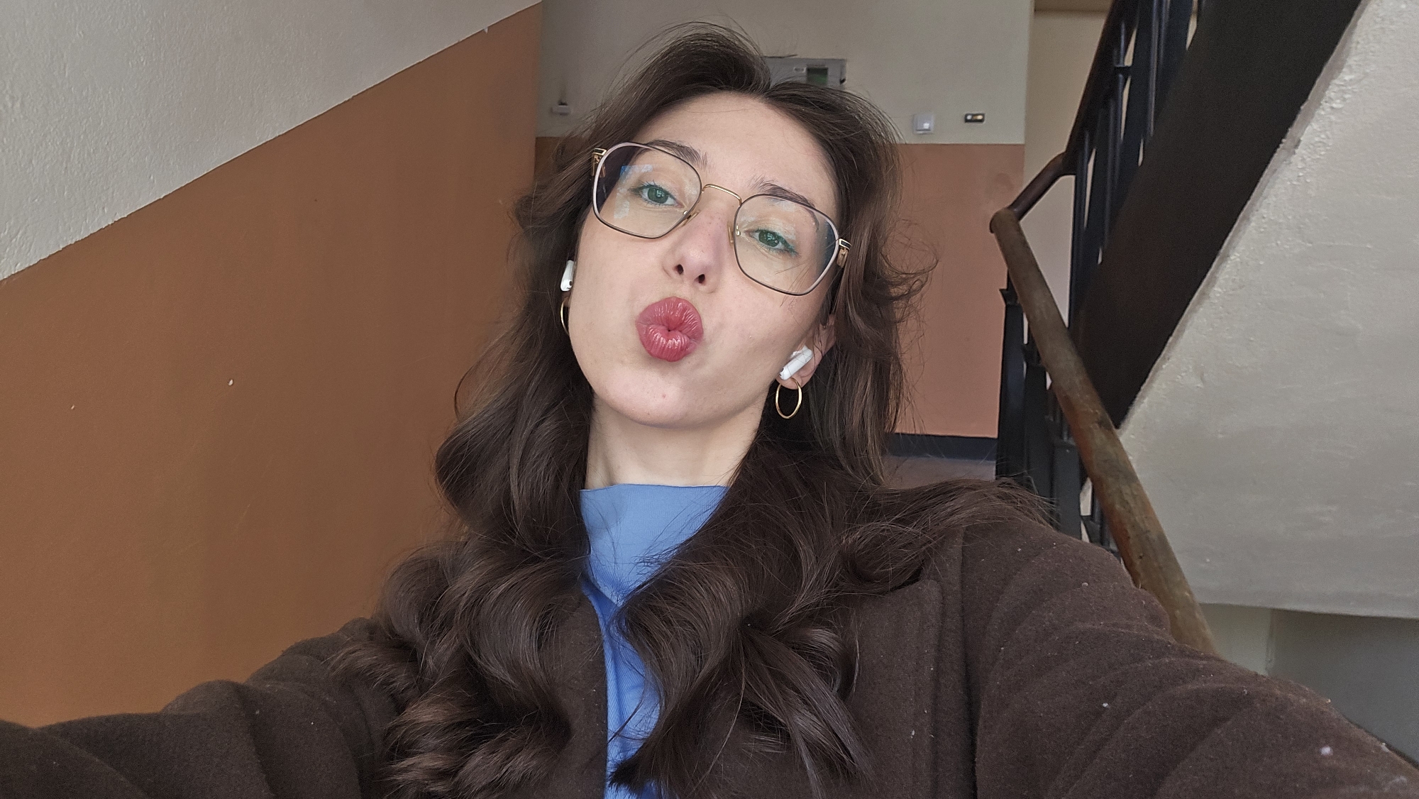 A woman is taking a selfie in what appears to be a stairwell. She is wearing glasses, gold hoop earrings, and white wireless earbuds. She has long, wavy brown hair, a blue top, and a brown coat. She is puckering her lips for the selfie. The background includes an orange wall, a staircase with dark metal railings and a wooden handrail, and a white wall. Based on visual context, it is not possible to precisely determine the location in the city of Stara Zagora, Bulgaria.