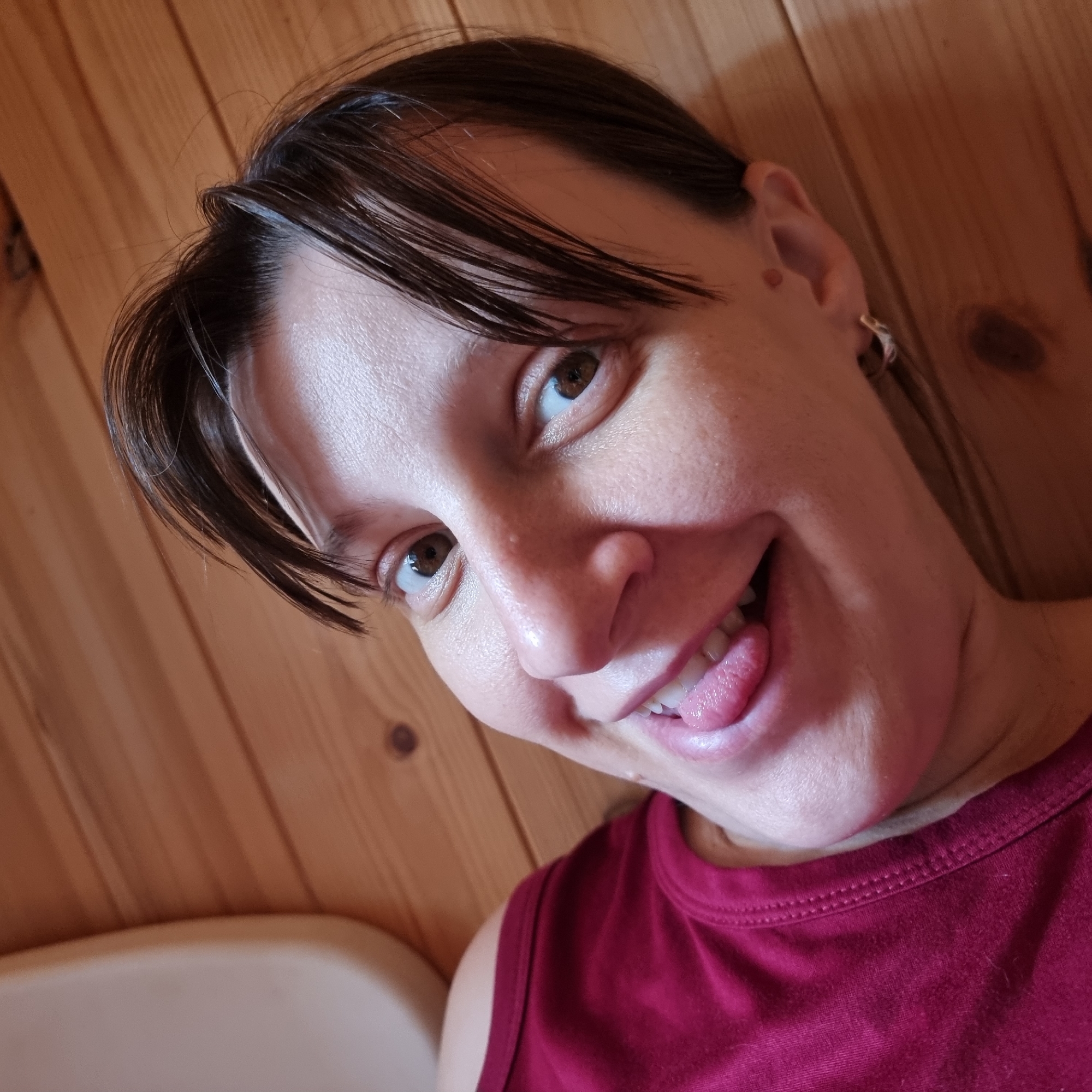 A woman is photographed close-up, with her head tilted and her tongue sticking out. She is looking towards the camera with a playful expression. Her dark hair is pulled back, with a few strands falling forward. She is wearing a maroon tank top. The background is a wooden wall, likely interior paneling, with visible grain and knot details. A white object, possibly a bathtub or sink, is partially visible in the lower left corner. The lighting is soft and natural. The image was taken indoors, likely in a home, in Dunnae-myeon, South Korea.
