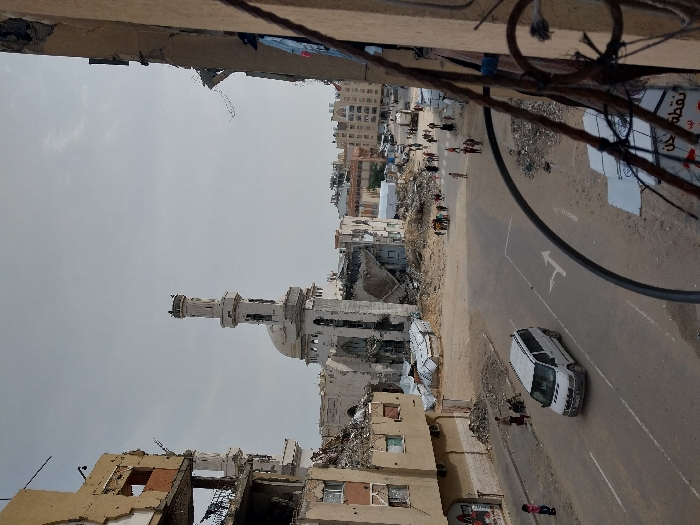 An elevated viewpoint photograph captures a severely damaged urban environment in Khan Yunis, Palestinian Territory, under an overcast sky. Central to the mid-ground stands a prominent mosque, light-colored, featuring a minaret and dome, both exhibiting significant structural damage with large sections of walls missing and rubble visible at its base. To the left, a multi-story building appears heavily damaged, with exposed rebar, collapsed floors, and significant portions of its facade missing. The immediate foreground on the right shows the edge of a damaged building, from which the image is taken, with twisted metal and electrical cables hanging.
A paved road with a white dashed center line and a painted white right-turn arrow runs diagonally across the lower half of the scene. A white minivan travels on this road, moving away from the viewer. The ground surrounding the road and damaged structures is covered with extensive piles of rubble, dirt, and debris. Several temporary shelters constructed from corrugated metal are visible among the debris near the mosque and damaged buildings.
Numerous individuals are present: a child in a pink shirt stands on the left sidewalk near the damaged building, while further along, a small group of people is gathered. A larger congregation of pedestrians, comprising adults and children, is seen walking and standing in the open, debris-filled areas near the damaged mosque.