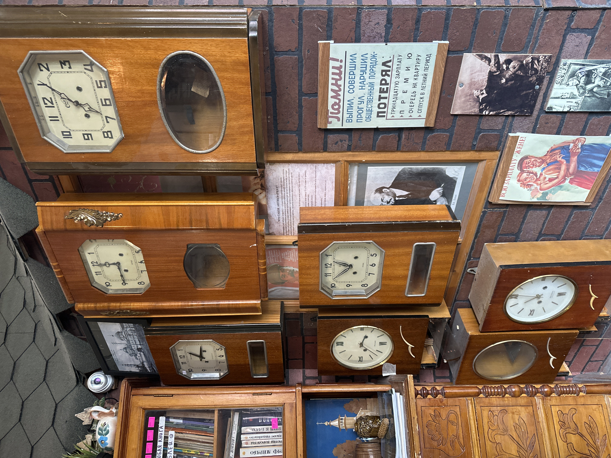 The image depicts a section of a wall cluttered with various objects, likely in Kazan, Russia. Several vintage wooden clocks are arranged vertically, each with different face designs and housing styles. To the right of the clocks are framed pictures and posters on a brick wall. One poster reads "Remember! Missed, committed Skipping, disturbed Public order - Lost," followed by a list of losses. Below the clocks and posters are more bookshelves and cabinets filled with books and decorative items, adding to the cluttered ambiance. The scene suggests a display in an antique store or a collector's cluttered space.