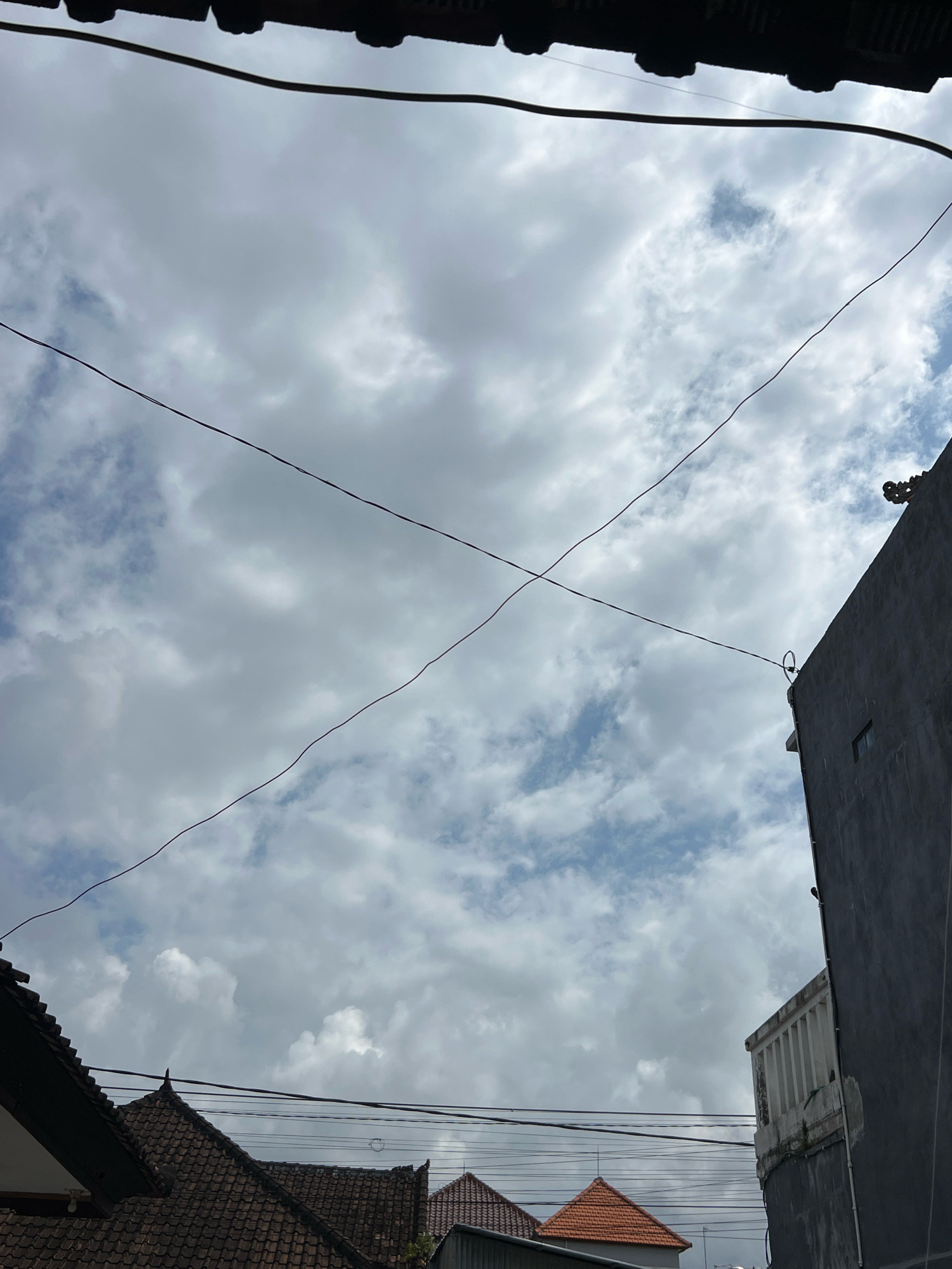 The image shows a view of the sky above a residential area, likely in Denpasar, Indonesia, characterized by overcast skies with scattered clouds and visible utility wires. The foreground features the dark, tiled roofs of traditional Indonesian houses, including one with a prominent, stepped roofline. To the right, a modern, gray concrete building with a white, balustraded balcony rises, partially obscuring the sky. The scene is captured from a low-angle perspective, looking upwards. There are no people visible in the frame. The overall impression is of a cloudy day in a densely built urban environment.