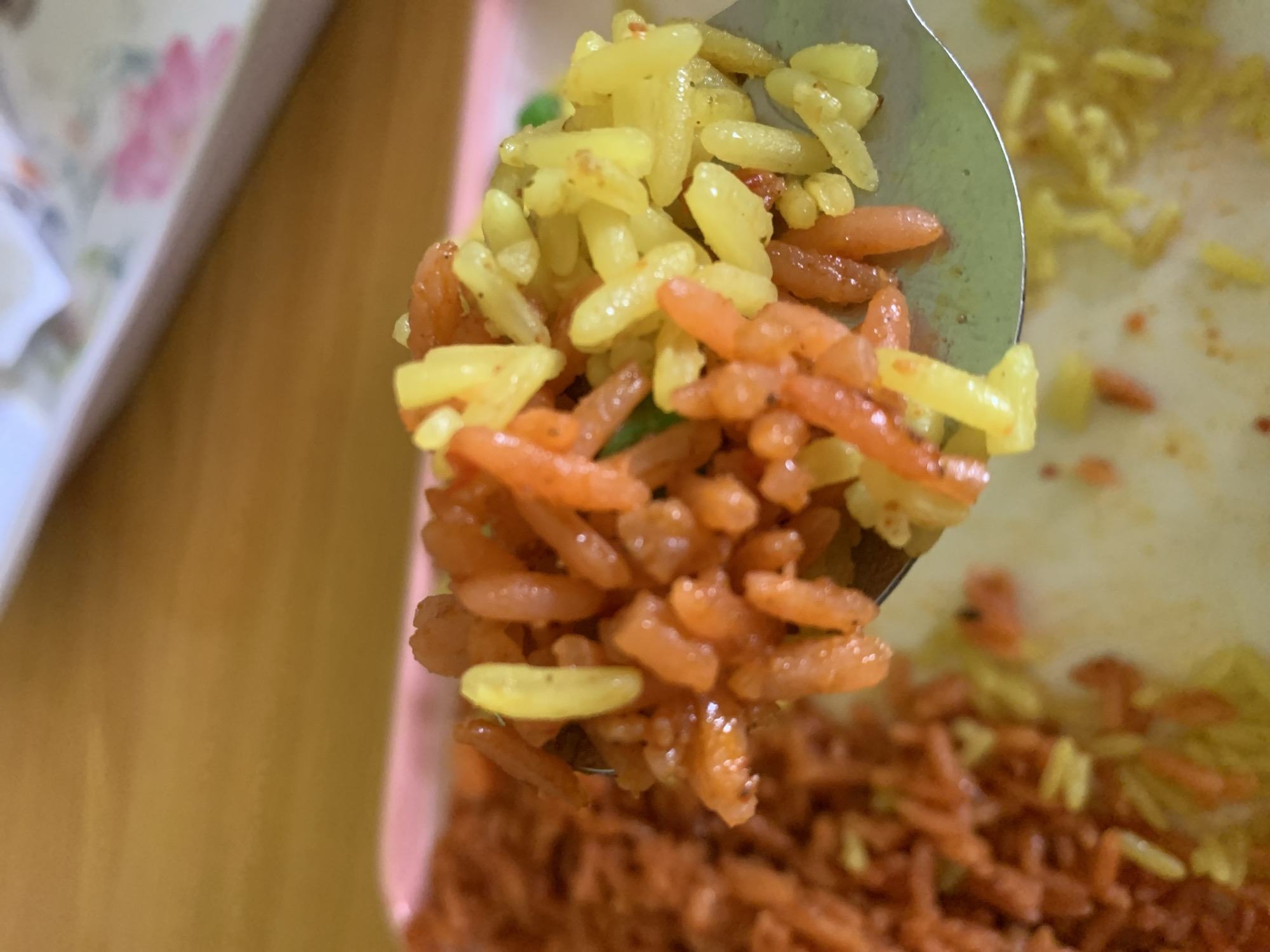 The image displays a close-up, slightly elevated perspective of a metallic spoon holding a portion of cooked multi-colored rice. The rice on the spoon consists of approximately equal quantities of orange-red and yellow elongated grains, with a few small green specks, consistent with peas or finely chopped vegetables, interspersed within. The rice grains appear separated and distinct. In the immediate background, a larger quantity of similar orange-red and yellow rice is visible on a light-colored, possibly pale pink, rectangular serving tray. The surface directly beneath the tray is a light brown wooden table. The upper right background reveals an out-of-focus white object exhibiting faint pink floral patterns. No human subjects or direct interactions are visible. The scene is located in New Stadium, Nigeria.
