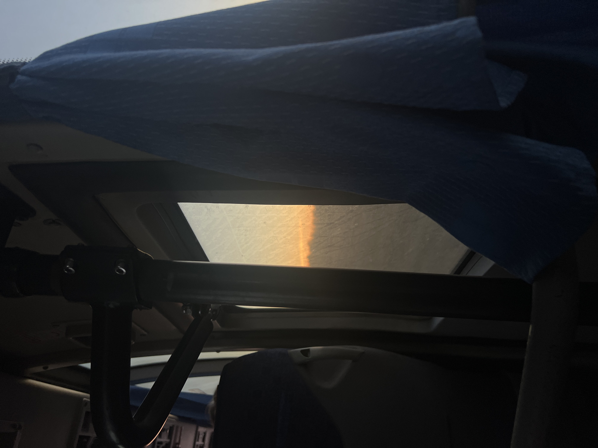 The image depicts an interior view looking upwards through a skylight. The skylight appears partially covered by a blue cloth. A black metal frame and support structure are visible in the foreground. The lighting outside is bright, possibly indicating sunrise or sunset. The image does not contain identifiable people, subjects, or actions. The location cannot be determined from the visual information provided.