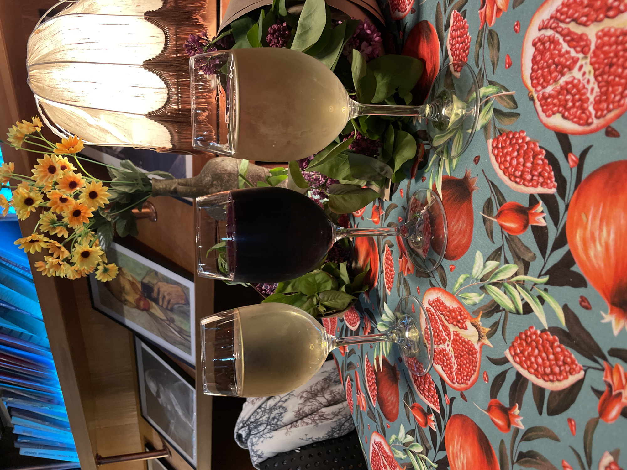 The image shows three wine glasses arranged on a table with a pomegranate-patterned tablecloth. The glasses contain a light-colored wine, red wine, and another light-colored wine. The glasses are positioned between floral arrangements, including sunflowers and greenery. In the background, there's a lit lamp with a decorative shade, artwork on the wall, and what appears to be a stack of blue objects. The location is likely Tbilisi, Georgia.