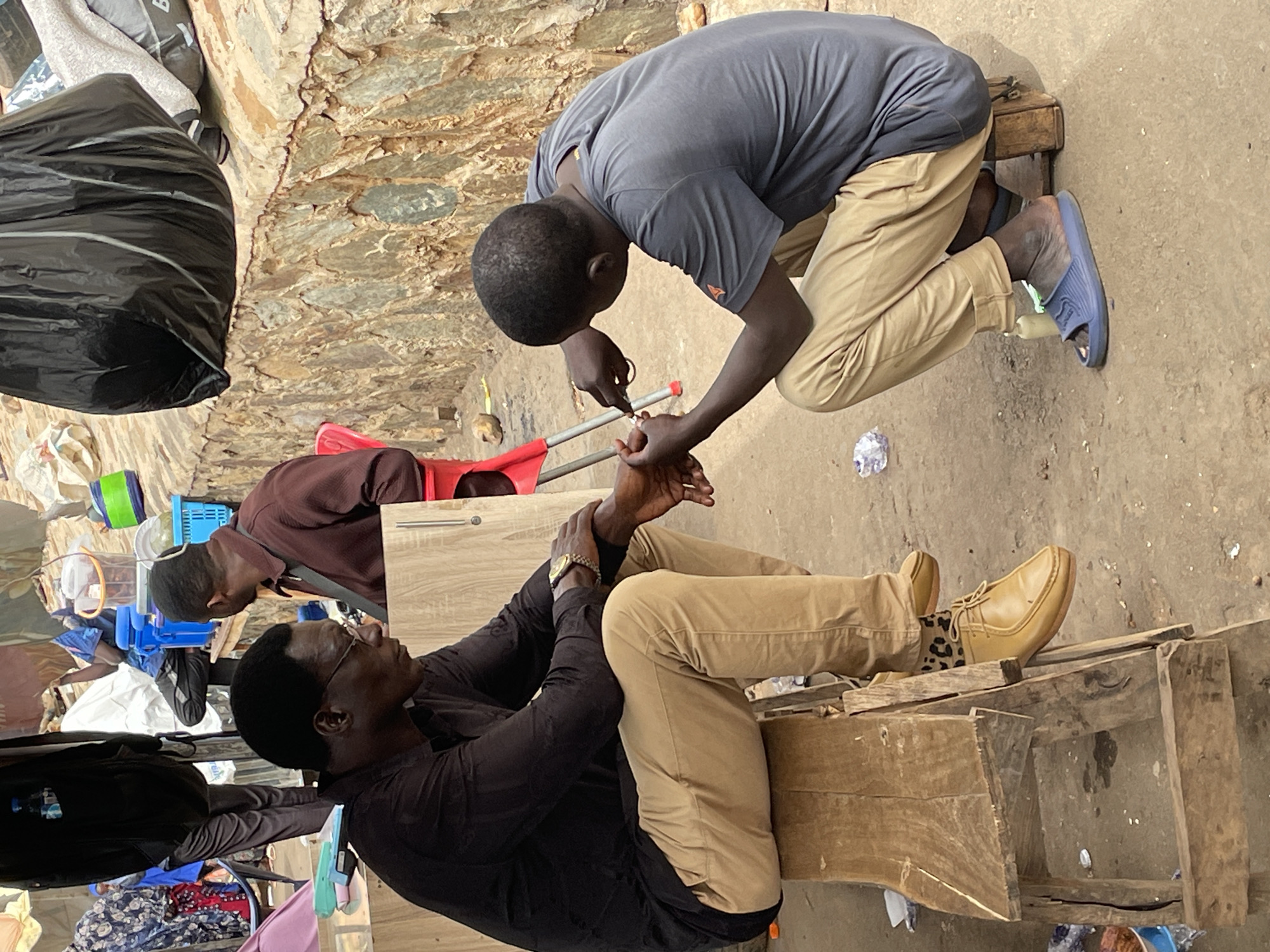 In Bukuru, Nigeria, two men are interacting in an outdoor setting. One man is kneeling and attending to the hands of the other, who is seated on a wooden structure. The kneeling man wears a grey shirt and khaki pants, with blue sandals nearby. He appears to be using a tool to examine or work on the seated man's hand. The seated man is dressed in a black shirt and khaki pants, wearing glasses and a watch. He's sitting on what appears to be a crude, wooden bench or platform and has a tan shoe on one foot. The background shows a mix of urban debris, items under tarps, and various household items.