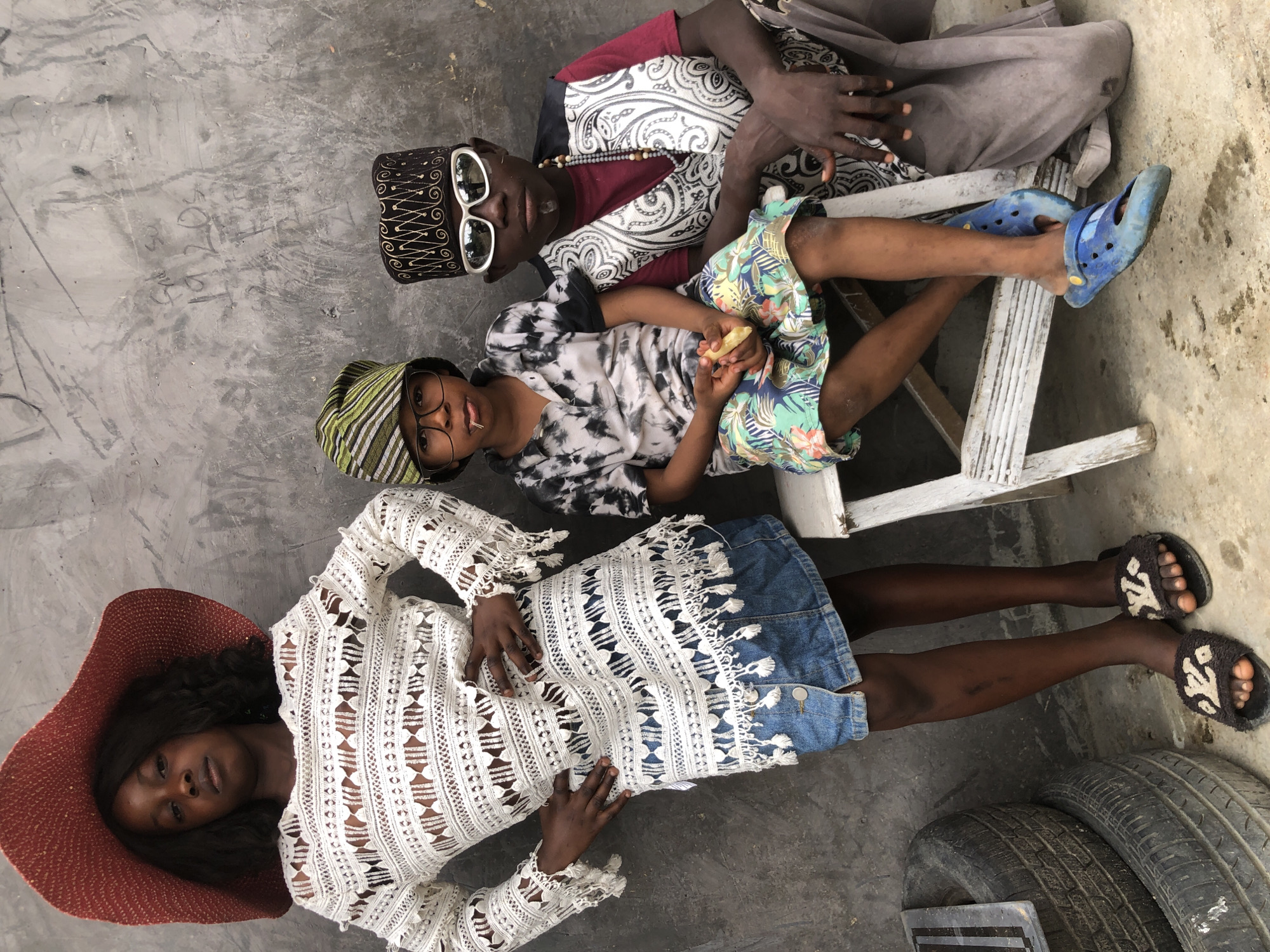 In Ado, Nigeria, three young people pose against a rough, grey concrete wall. On the left, a girl stands wearing a large, red sun hat and a white, intricately patterned, long-sleeved crochet dress over a denim skirt. She has her hands on her hips. To her right, seated on a white, slatted wooden stool, are two boys. The boy on the left wears a green and white striped cap, glasses, and a tie-dye t-shirt with a colorful patterned skirt. He holds a piece of food in his hand and has a piece of food in his mouth. The boy on the right wears a black and gold patterned cap, white sunglasses, a red shirt, and a black and white patterned vest. He has a beaded necklace and is holding a piece of food. Behind the children, the wall is marked with graffiti, including the numbers "1920s". To the left of the girl, a stack of tires is visible. The ground is concrete.
