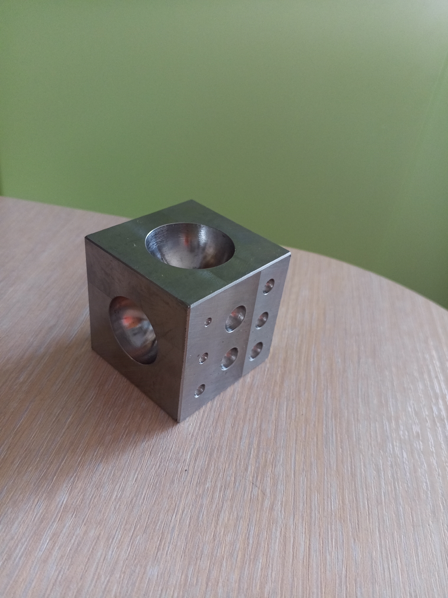 A metallic cube with multiple holes and indentations sits on a light-colored wooden table. The cube has a large circular opening on its top face and another on its left face. The front face of the cube features several smaller circular indentations. Behind the table, a plain light green wall is visible. The image is taken from a medium-angle perspective, focusing on the cube and the table surface. There are no people present in the image. The scene does not provide any specific geographical indicators to determine its location within Vilyuysk, Russia.