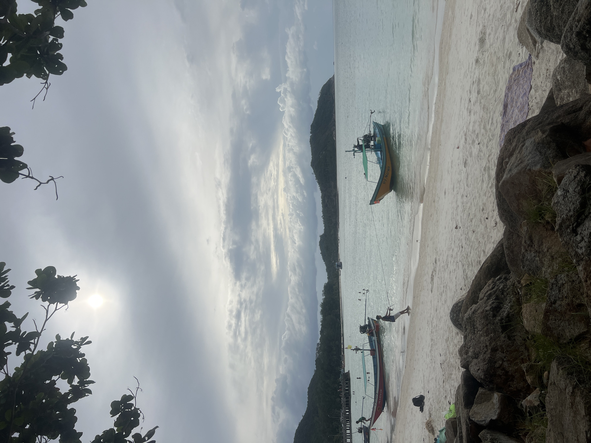 The image depicts a beach scene in the Ko Pha-ngan District, Thailand, under an overcast sky with the sun faintly visible through the clouds. In the foreground, large rocks covered with sparse grass line the edge of a sandy beach. A purple and white patterned towel is laid out on the sand. In the middle ground, the shallow, clear turquoise water laps at the shore. Two longtail boats are anchored in the water. One boat, primarily red with white and blue accents and the name "99 MARWIN" visible, is closer to shore. A person in a dark shirt and white shorts is standing in the water near this boat, holding a fishing rod and appearing to cast a line. Another person in a green shirt is in the red boat. A second longtail boat, painted yellow and blue with a green canopy, floats further out in the water. The background features a line of lush, green hills and islands, with a distant pier visible on the left. The overall atmosphere is tranquil and suggests a cloudy day at a tropical beach.