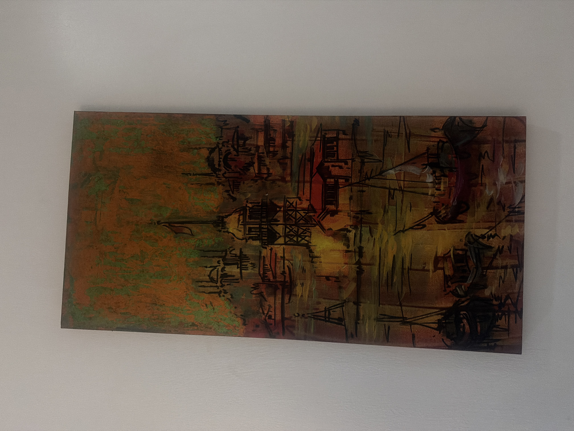 The image shows a framed abstract painting mounted on a white wall. The painting features a mix of brown, green, orange, and yellow hues with black linear details that suggest an urban or industrial landscape. The painting's style leans towards abstract expressionism. The location is not discernible from the image and there is no information to place it in Iddo Tudunwada, Nigeria.