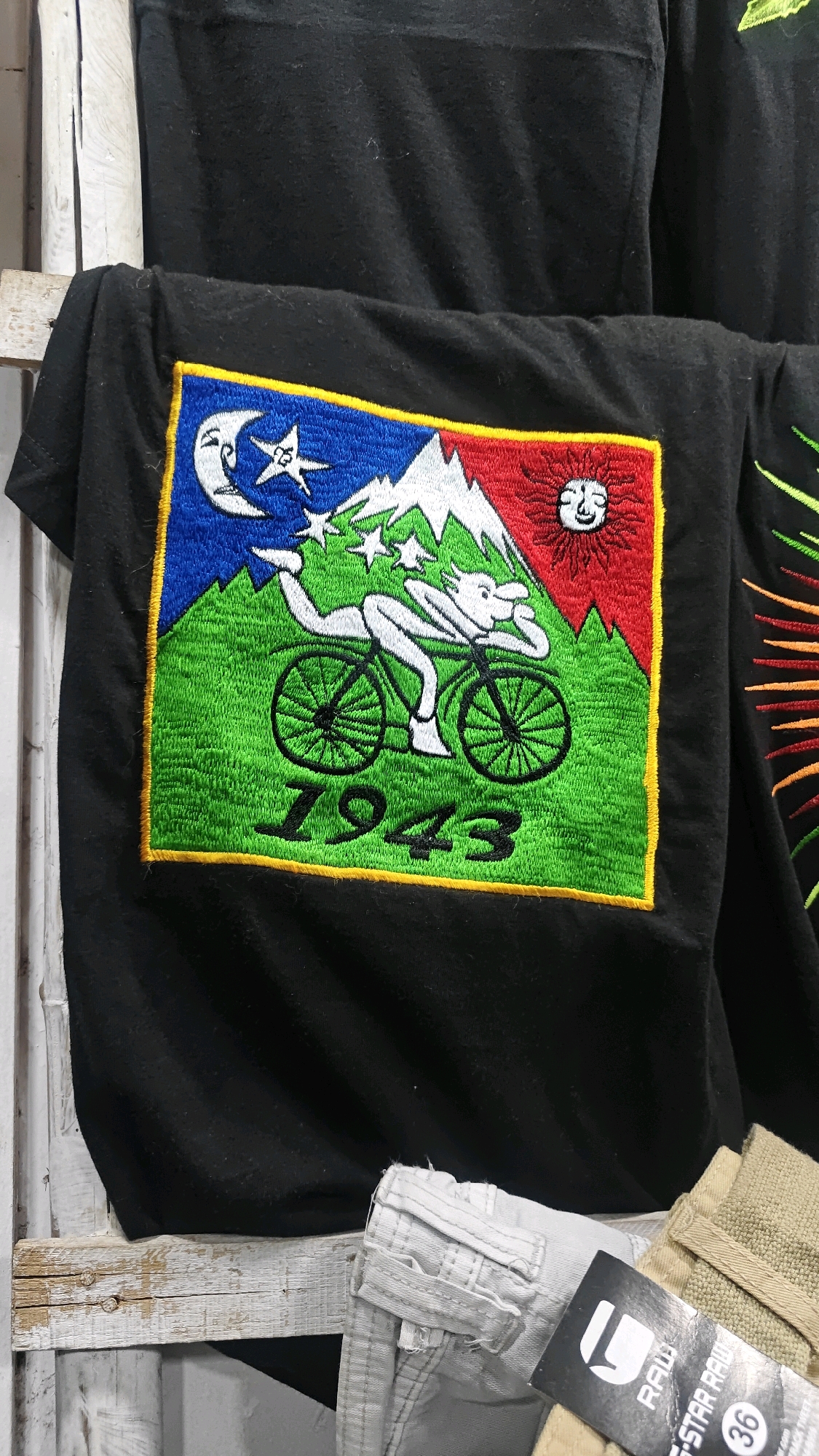 A black t-shirt with a vibrant, embroidered graphic is displayed on a white wooden structure. The graphic depicts a stylized figure riding a bicycle through a landscape. The scene includes a crescent moon and stars on the left, mountains in the center, and a sun with a face on the right. The word "1943" is embroidered in black text at the bottom of the graphic. Below the t-shirt, a pair of light-colored pants and a beige fabric item are visible, along with a tag that reads "G-STAR RAW 36". This scene is located in Anjuna, India.