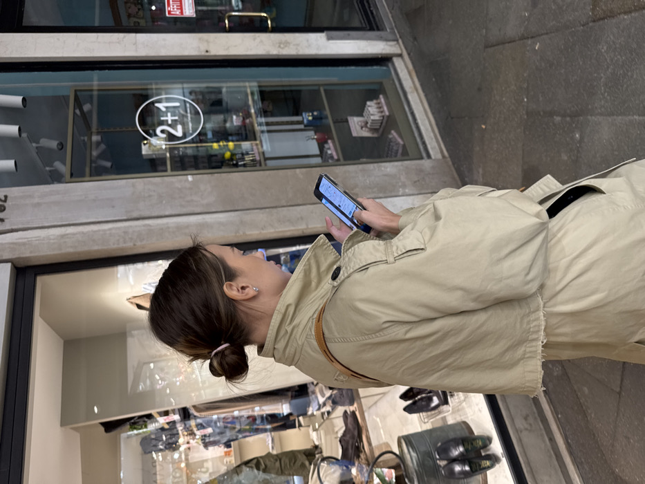 A single adult female is depicted in profile, standing on a grey stone-paved sidewalk in a commercial area of Venice, Italy. She wears a light beige trench coat, with a brown strap, possibly from a bag, visible over her right shoulder. Her medium brown hair is styled in a messy bun secured with a light pink elastic, and she wears a small silver hoop earring in her left ear. She is actively engaged with a dark-colored smartphone, holding it with both hands and looking down at its illuminated screen.
Behind her are two storefronts with large glass display windows. The left window showcases clothing and shoes, including black loafers and boots, with a green metal container at the bottom of the display. The right storefront, separated by a light-colored stone pillar, features interior shelving with various small products. A circular white decal with "2+1" is visible on the glass of this window. Above the right storefront, the building number "782" is visible. An "AFFITTO" (For Rent) sign, white with red lettering, is affixed to the frame of the right storefront's door. The street pavement is composed of large, irregularly shaped grey stone slabs.