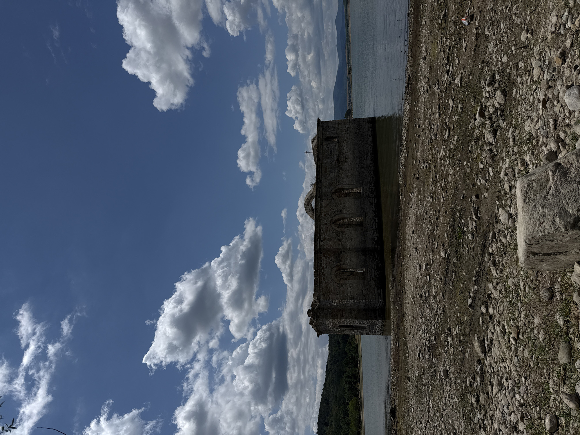 The image shows the partially submerged ruins of a stone church on the shore of a lake. The church, located in Panicherevo, Bulgaria, is made of weathered stone blocks and has arched windows. Part of the roof is missing, revealing the interior structure. The church is surrounded by water, with the shoreline composed of pebbles and dry earth. In the background, a green, tree-covered hill rises from the far side of the lake, with distant mountains visible on the horizon under a blue sky dotted with fluffy white clouds.