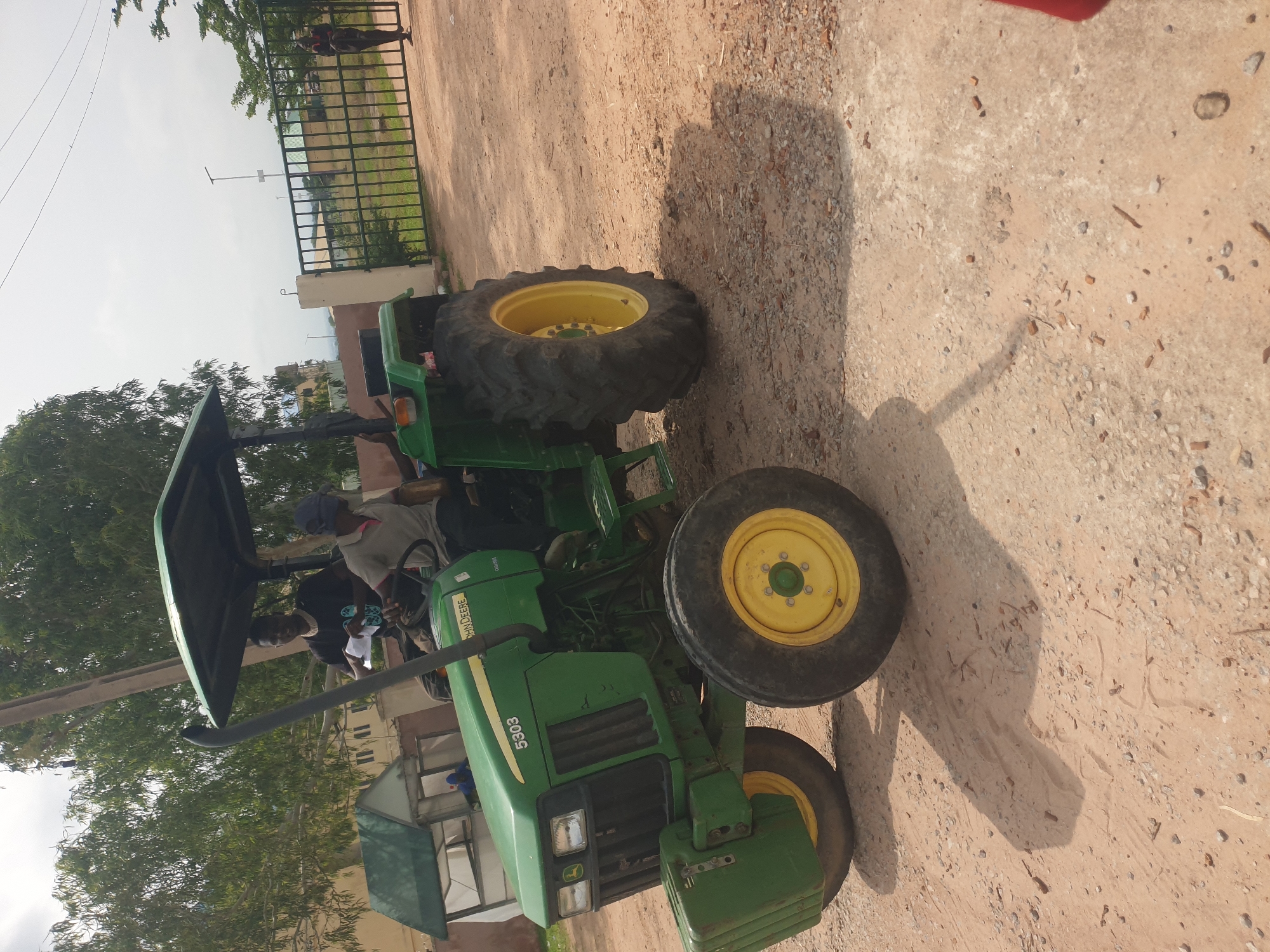 In Jalingo, Nigeria, a green John Deere 5303 tractor is parked on a dirt lot. Two individuals are seated on the tractor. The driver, wearing a grey shirt and a blue head covering, is partially obscured by the steering wheel. Behind him, another person in a black t-shirt and a necklace holds a white object, possibly paper. The tractor has a black canopy overhead and prominent yellow wheels. To the right of the tractor, a black metal gate with vertical bars is visible. Beyond the gate, a building with a tan facade and a green roof is situated behind a line of trees. A single individual in dark clothing stands near the gate. The ground is a mixture of sand and small stones, with shadows cast by the tractor and surrounding objects.