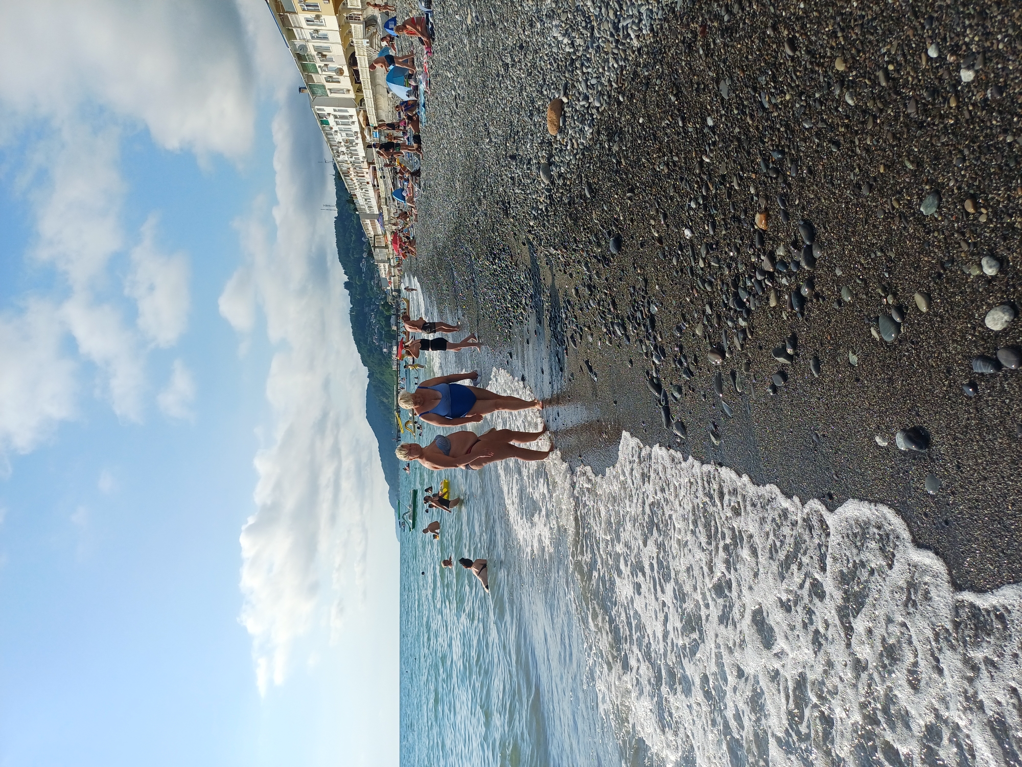 Two women are walking on a pebbled beach in Sochi, Russia. The woman on the left is wearing a dark bikini, and the woman on the right is wearing a blue swimsuit. They are walking towards the water, which is calm and has gentle waves. In the background, several other people are swimming and sunbathing. Behind the beach, there are buildings and lush green hills. The sky is blue with scattered clouds.